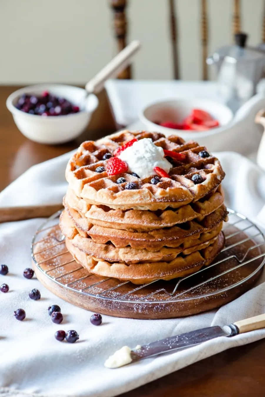 Waffle 桌布 壁紙 — 一疊高高的金黃色鬆餅，上面放滿了鮮奶油、草莓和藍莓，放在金屬架上，周圍散落著莓果，旁邊有一把奶油刀。