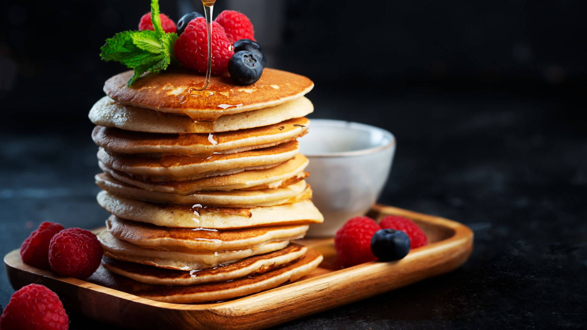 Pancakes 桌布 壁紙 — 一疊高高的金黃色鬆餅，淋上蜂蜜，搭配新鮮莓果和薄荷，放在木托盤上。