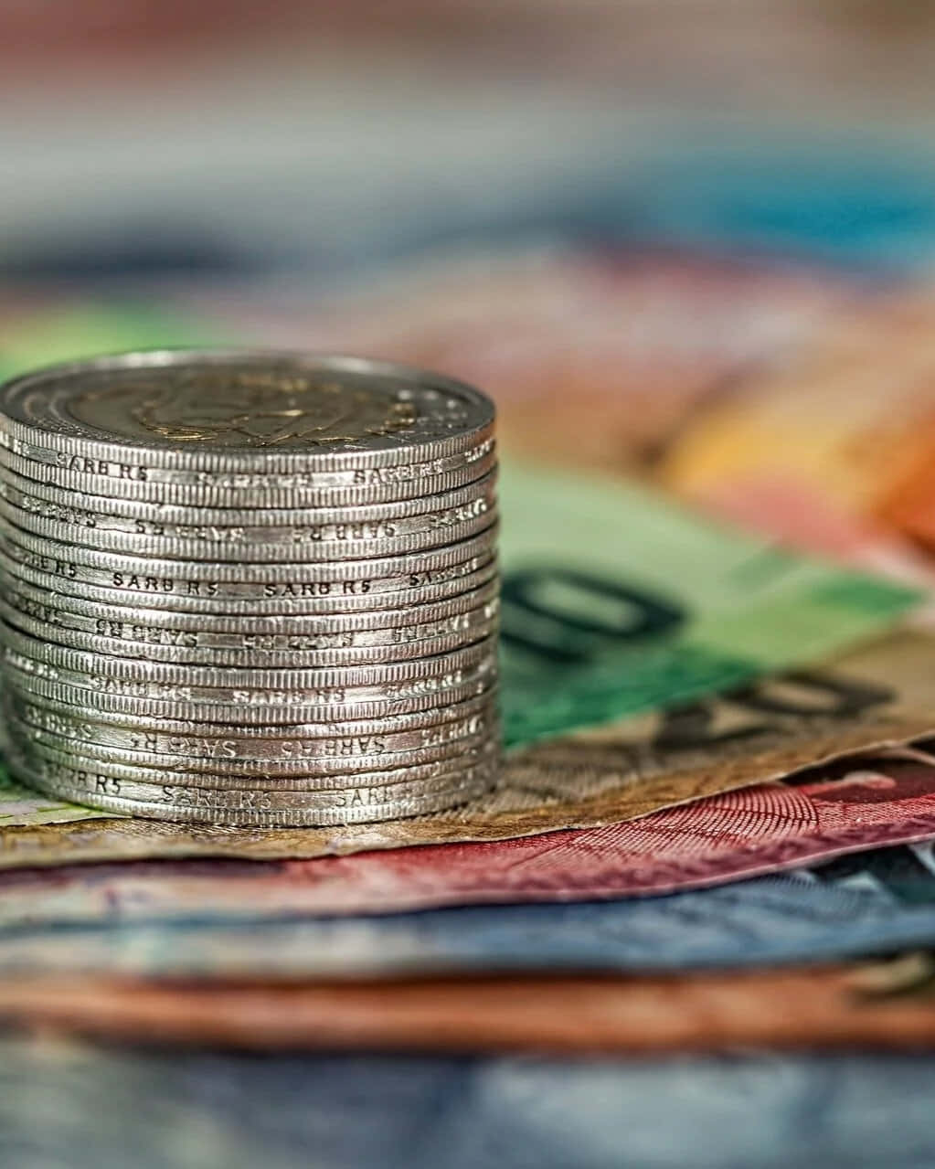 金錢 桌布 壁紙 — 一疊高高的銀幣精確地放在一堆色彩繽紛的鈔票上，背景模糊。