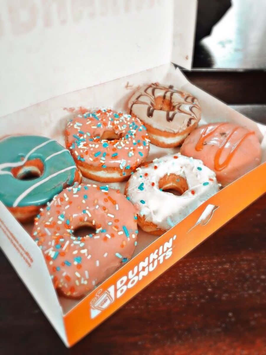 Dunkin Donuts 桌布 壁紙 — 一盒色彩繽紛的Dunkin' Donuts甜甜圈，有粉紅、藍和白色糖霜及彩針，放在木桌上。