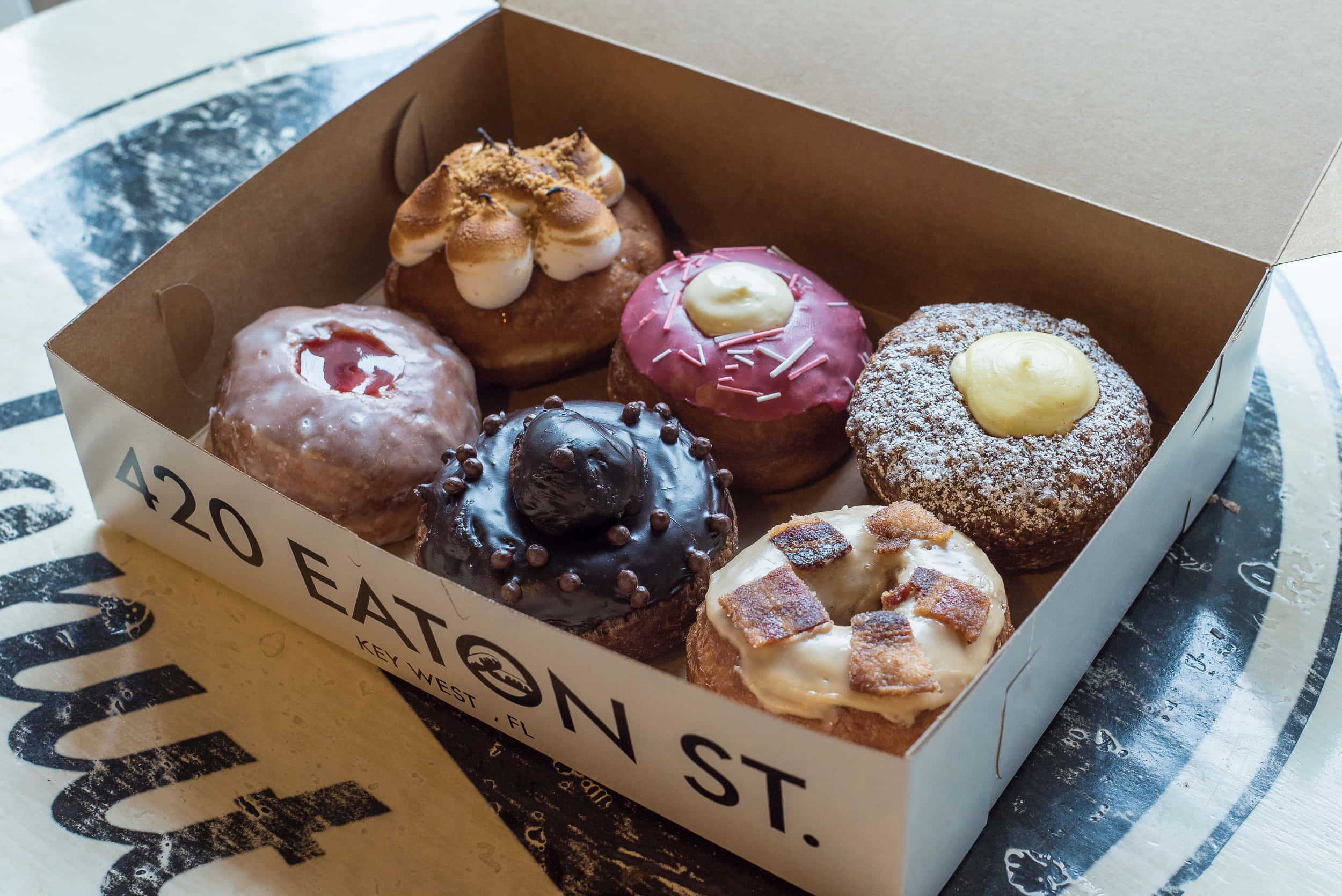 Glazed Donut 桌布 壁紙 — 一盒誘人的各式精選美食甜甜圈，配上獨特配料，放在圖案桌布上。