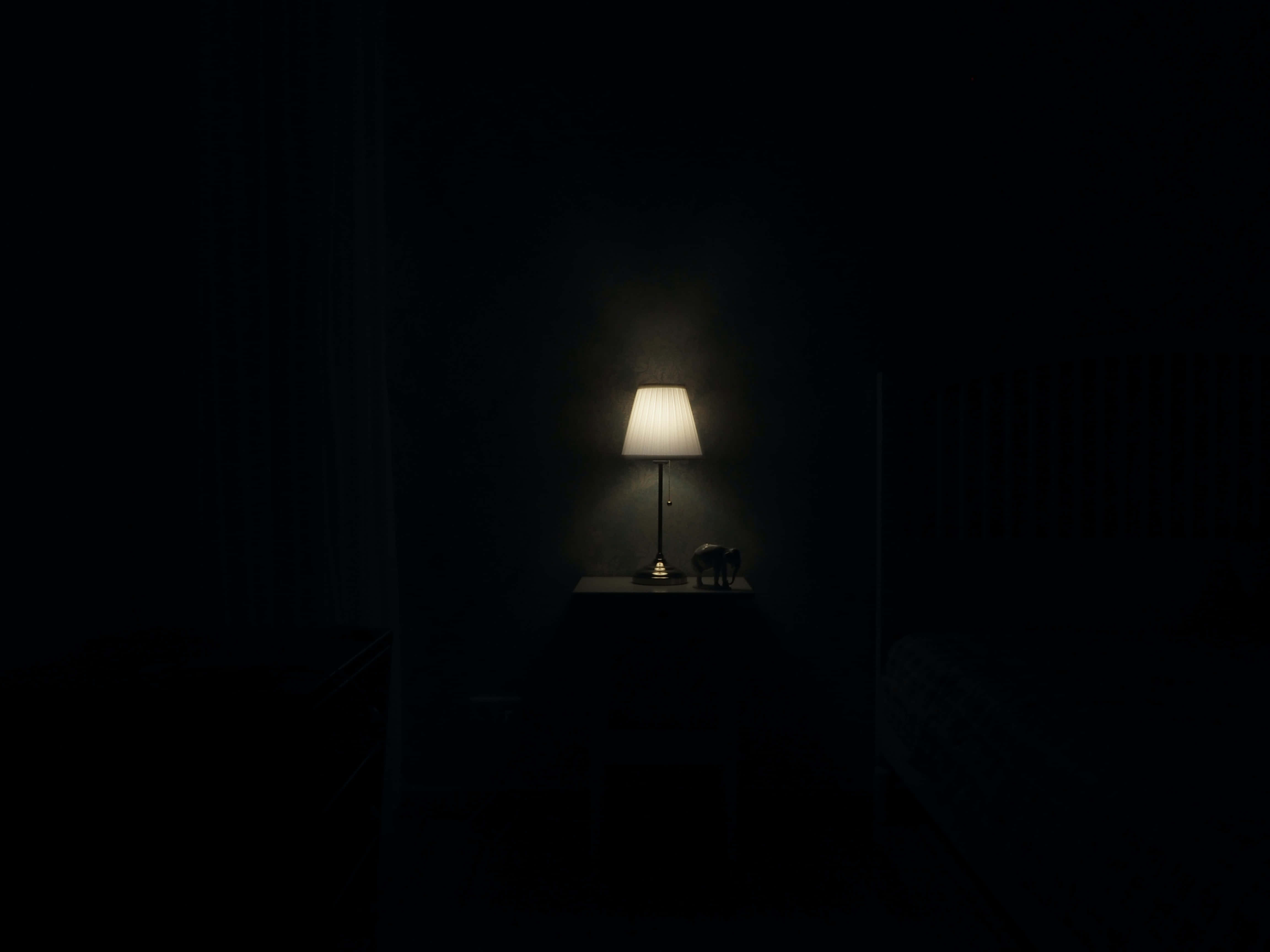 Dark Room 桌布 壁紙 — 一盞亮著的燈在漆黑的臥室中，在床頭櫃上投下溫暖的光芒。