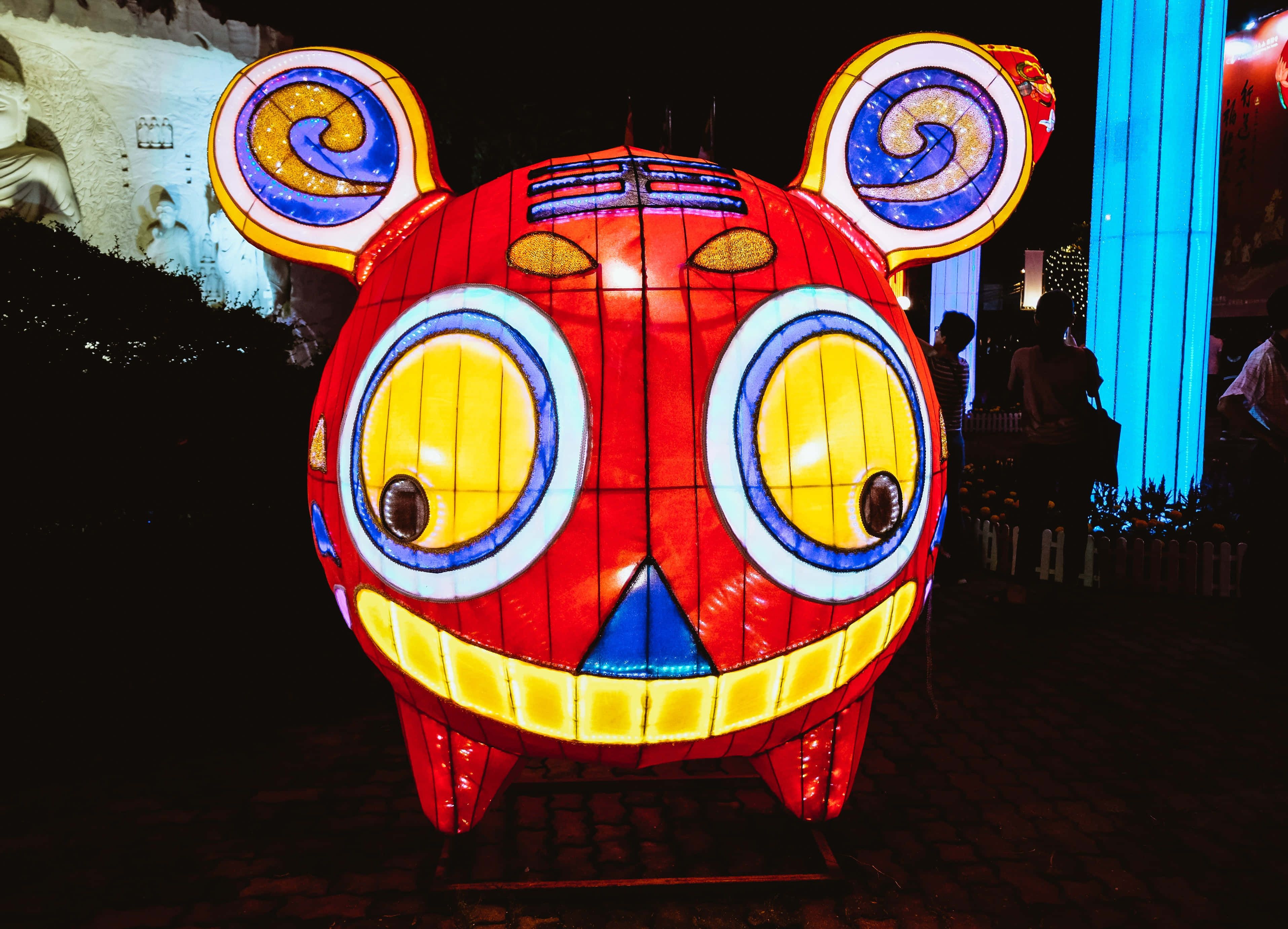 Festival 桌布 壁紙 — 一盞明亮照明的紅色卡通老虎燈籠，有黃色眼睛和螺旋耳朵，在夜晚的節日戶外環境中發光。