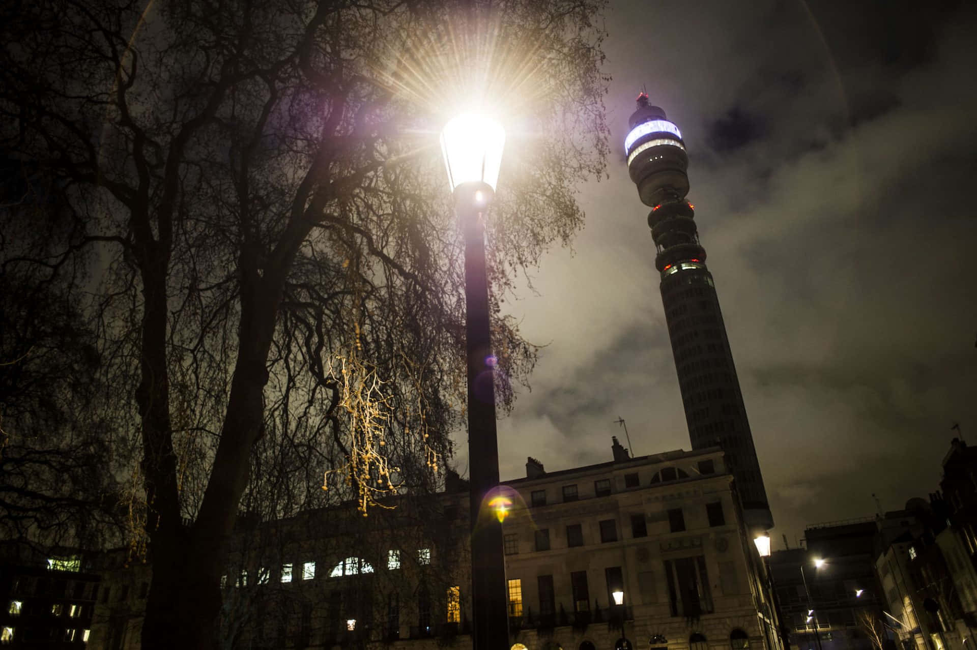 tower 桌布 壁紙 — 一盞明亮的街燈在陰雲密布的夜空中發光，勾勒出光禿樹木的輪廓，並照亮了倫敦標誌性的BT塔。