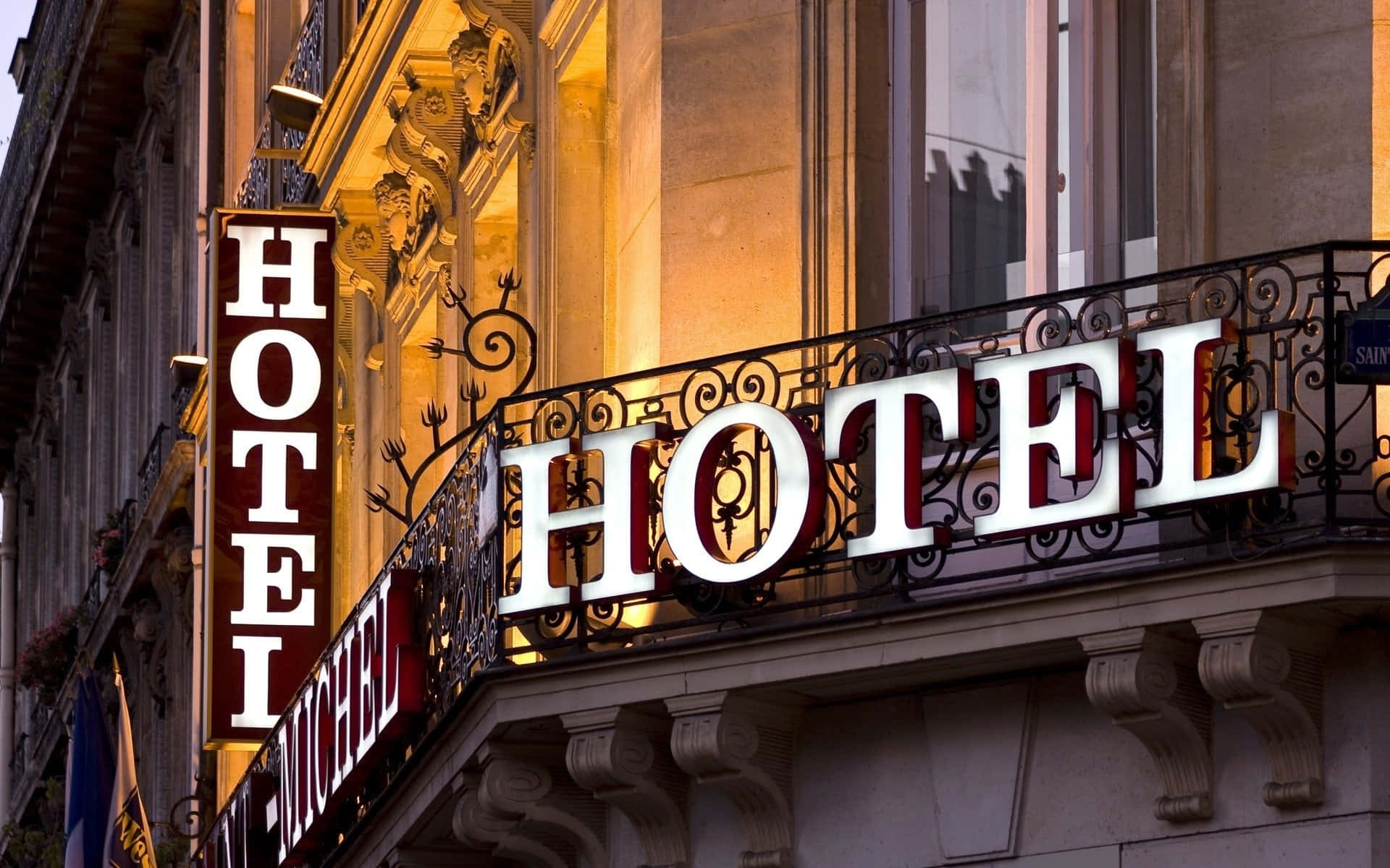 Hotel 桌布 壁紙 — 一盞溫暖照明的酒店招牌，帶有華麗的鐵藝裝飾，在黃昏時分映襯著經典歐洲建築的外牆。
