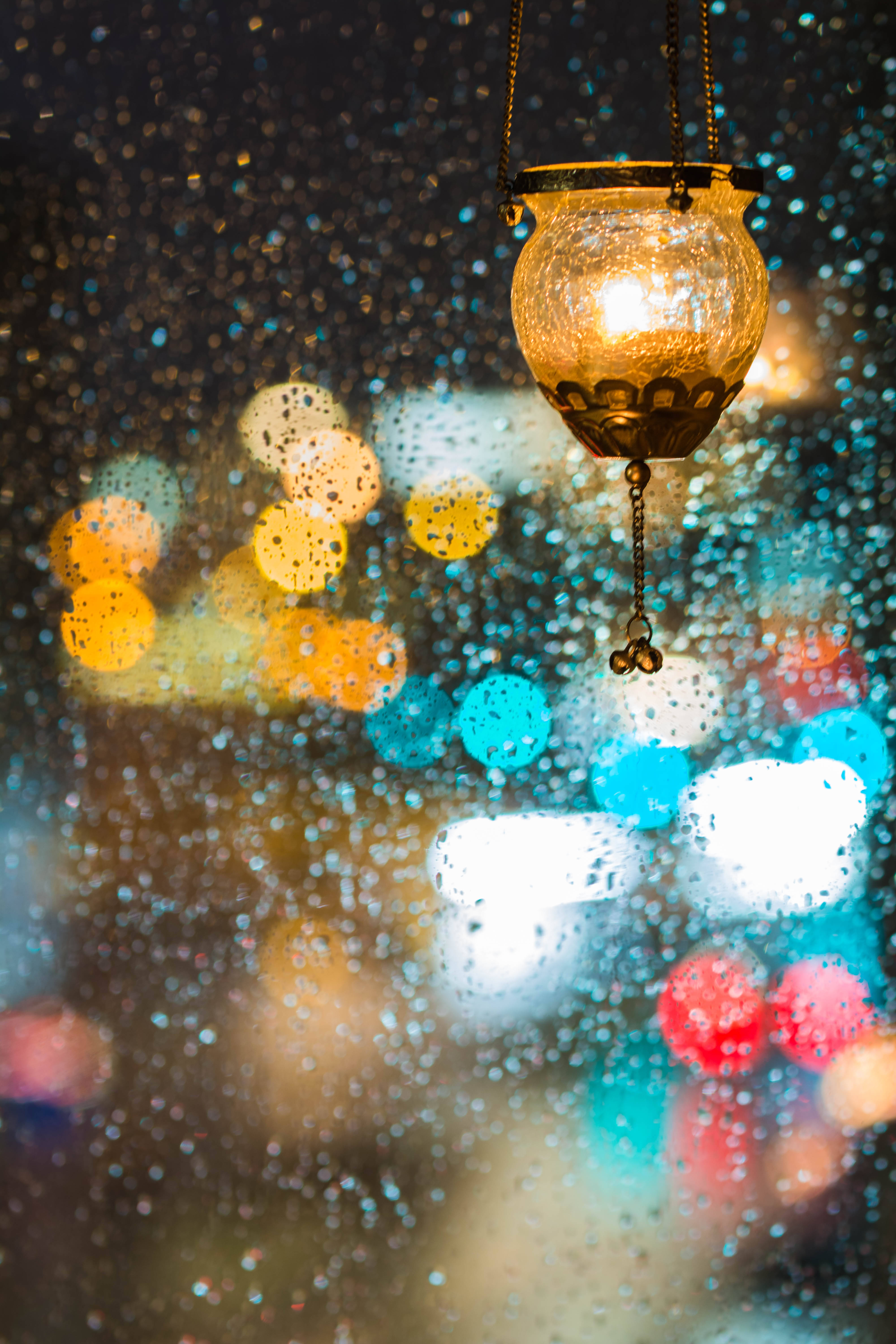 Bokeh 桌布 壁紙 — 一盞溫暖發光的燈籠懸掛在佈滿雨點的玻璃後，背景是夜晚充滿活力的散景城市景觀。