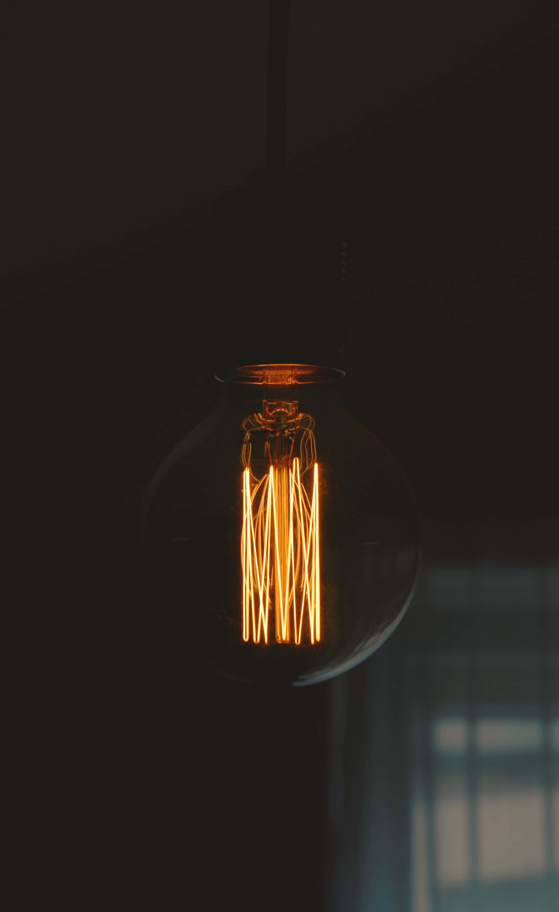 Light 桌布 壁紙 — 一盞發光的復古燈絲燈泡懸掛在黑暗中，透過玻璃外殼散發出溫暖的金色光芒。