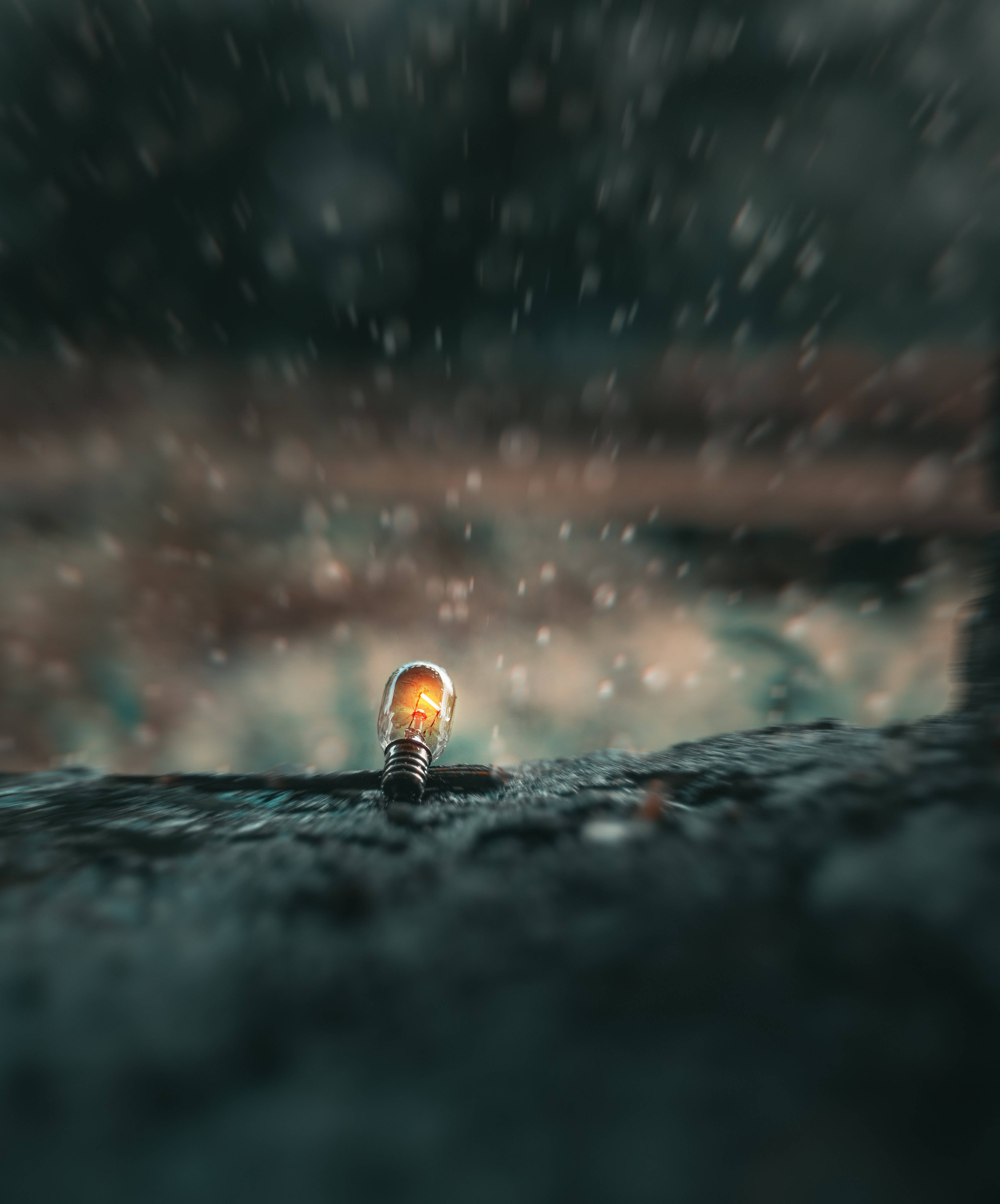 Focus 桌布 壁紙 — 一盞發光的燈泡在潮濕的表面上挑釁地閃爍，雨滴落在周圍，營造出陰鬱、充滿氛圍的場景。