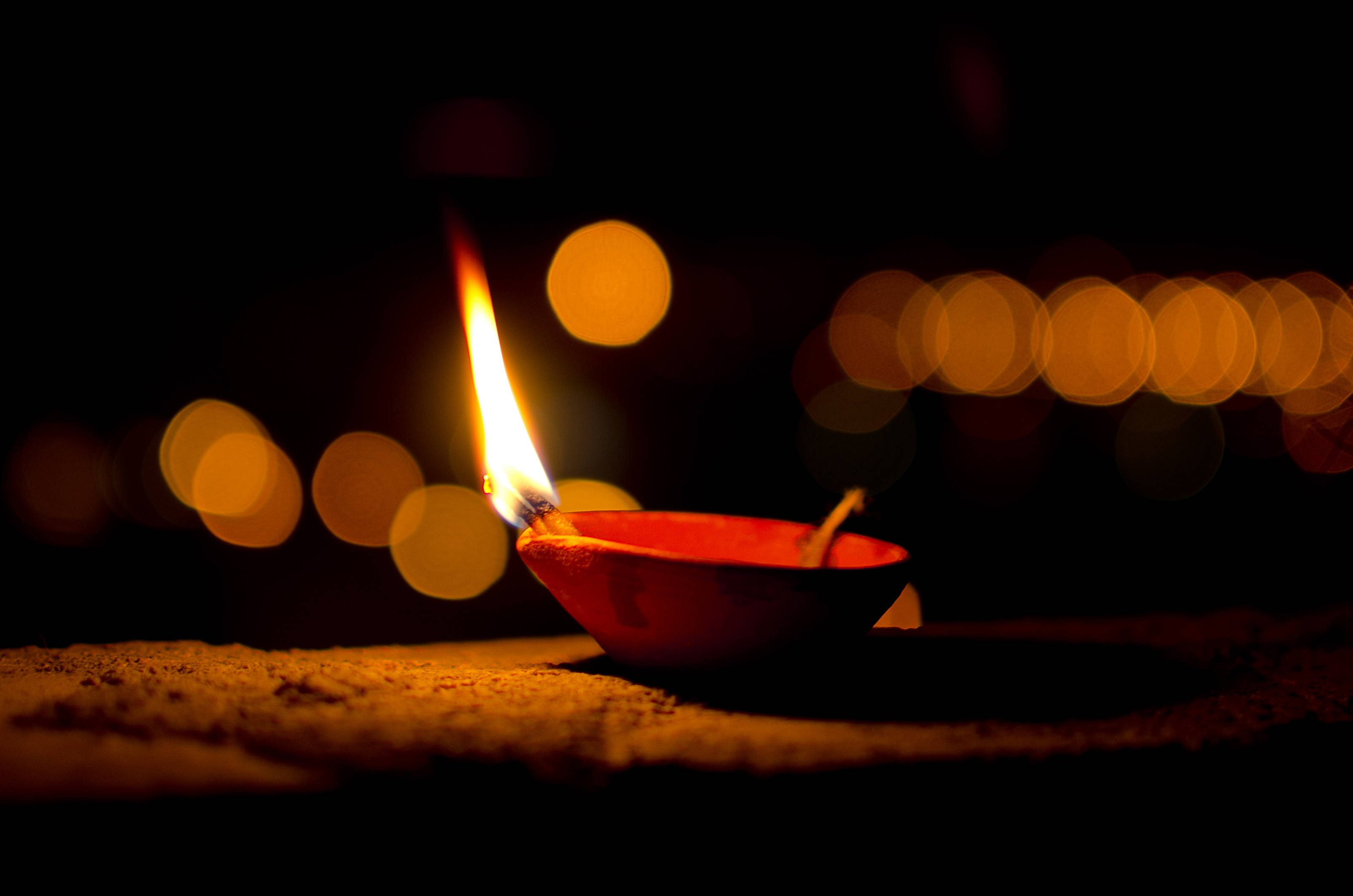 Happy Diwali 桌布 壁紙 — 一盞發光的紅色油燈在黑暗背景下，將溫暖的光線投射在沙地上，並帶有金色散景燈光。