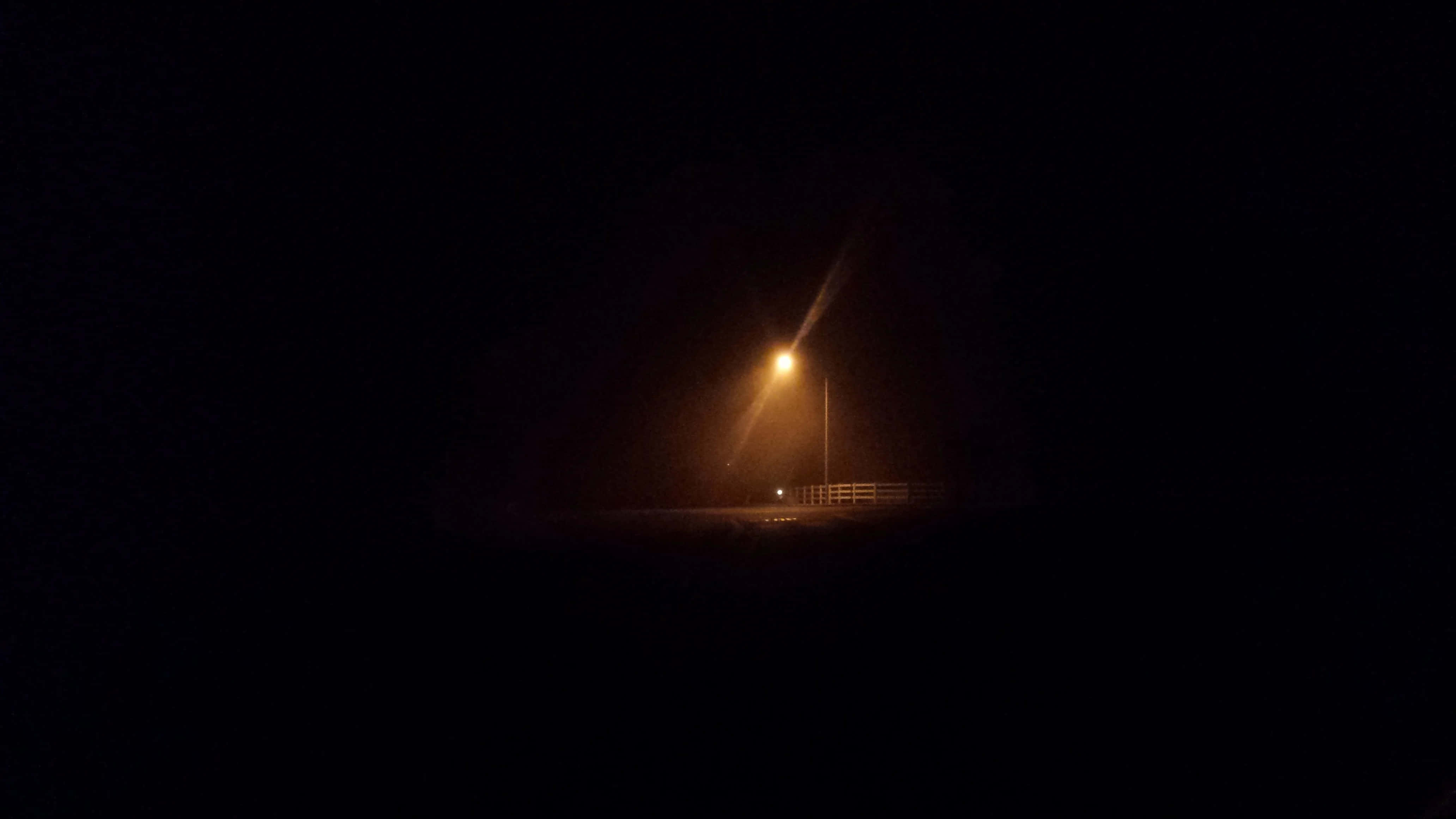 Dark Crossroad 桌布 壁紙 — 一盞發光的街燈刺破霧夜的黑暗，在遠處的圍欄和道路上投下溫暖的光環。