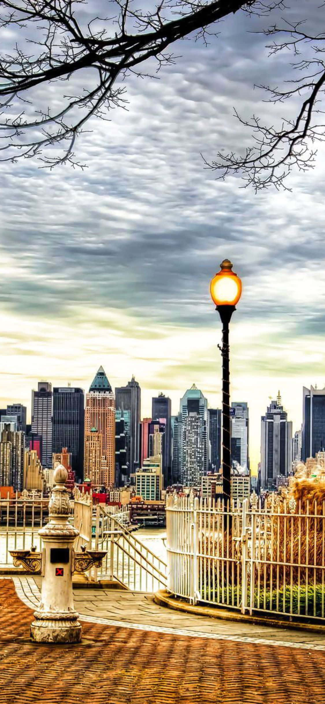 iPhone Xs Ny City 桌布 壁紙 — 一盞發光的街燈照亮了一條鵝卵石小徑，俯瞰著陰雲密布的天空下廣闊的城市天際線。