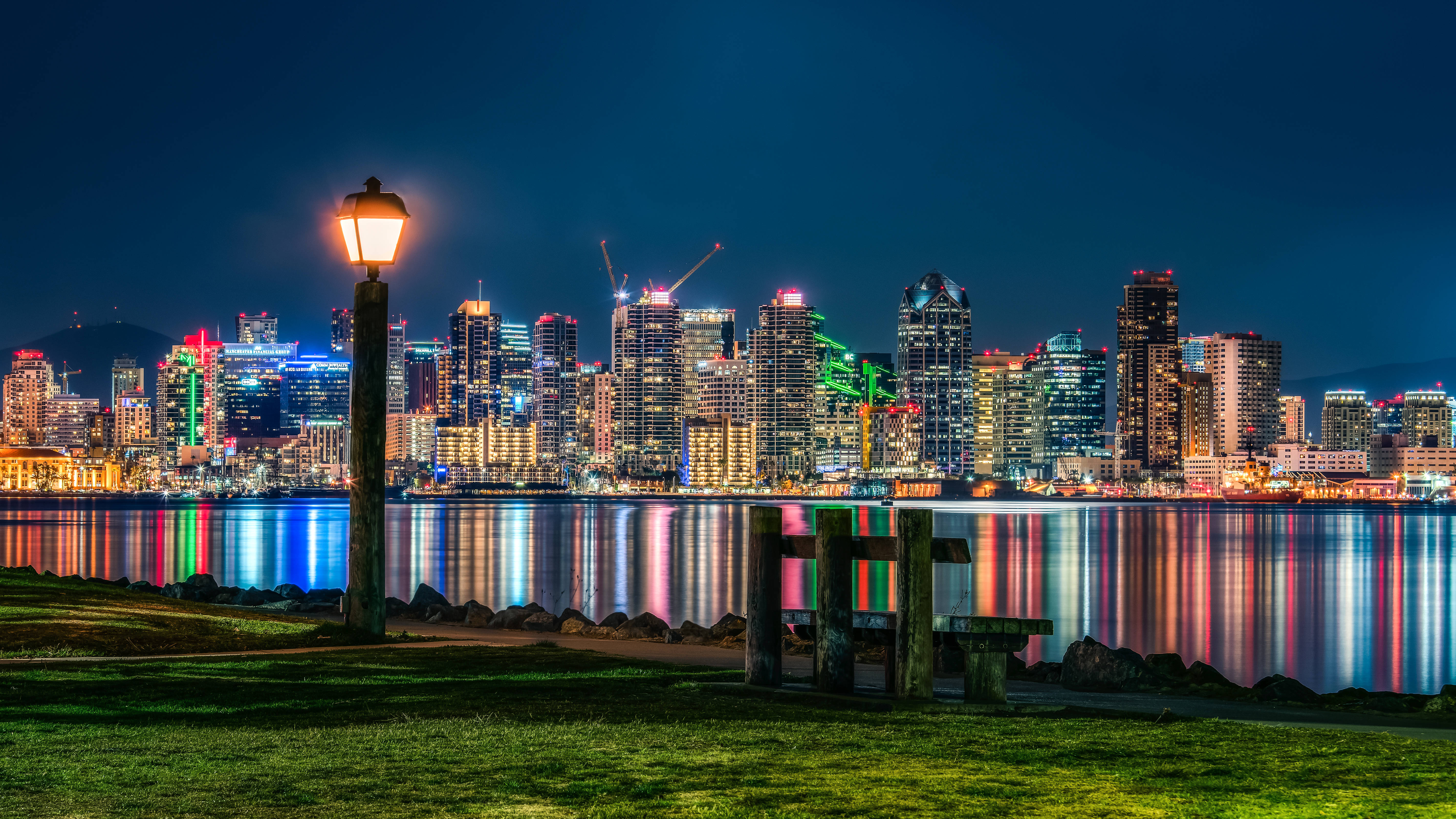 lights 桌布 壁紙 — 一盞發光的街燈照亮了夜晚的草地海濱，將繁華城市天際線的鮮豔霓虹燈光反射在水面上。
