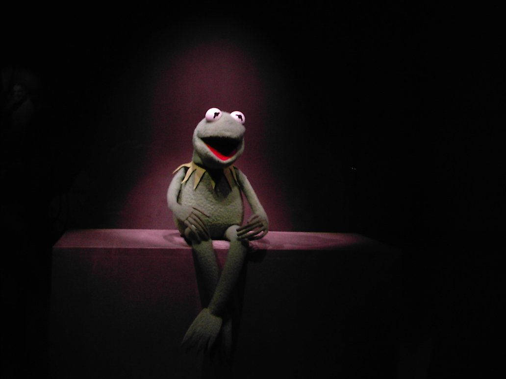kermit 桌布 壁紙 — 一盞聚光燈照亮了坐在黑暗劇院基座上的青蛙柯密特，他睜大的眼睛和張開的嘴巴表達了驚訝或喜悅。