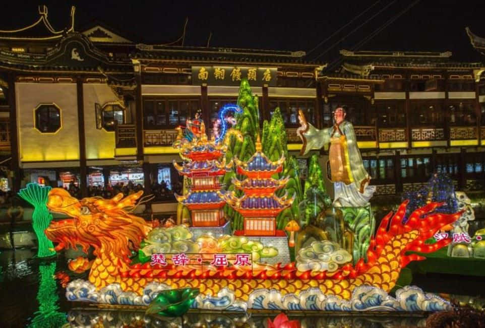 春節 桌布 壁紙 — 一盞色彩鮮豔的發光龍燈籠在夜間漂浮在水面上，周圍是傳統的中國建築和閃爍的節日裝飾。