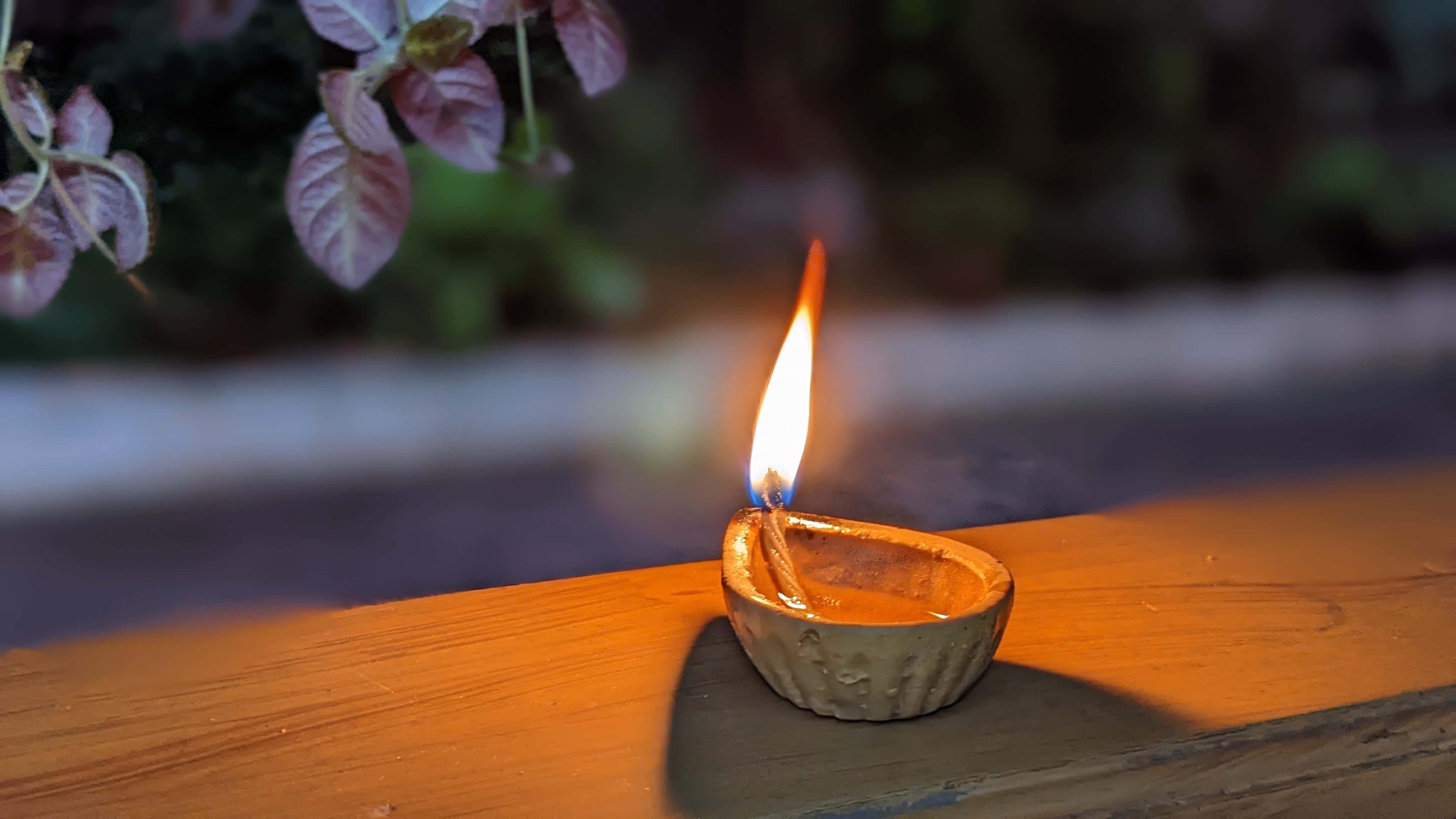 排燈節 桌布 壁紙 — 一盞點燃的油燈在木質表面上投射出溫暖的金色光芒，背景是模糊的紫色樹葉。