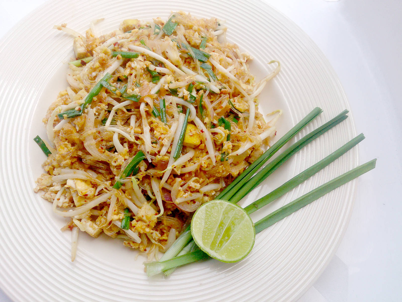 thai 桌布 壁紙 — 一盤充滿活力的泰式炒河粉，配上豆芽、青蔥和萊姆角，放在白色紋理盤上。
