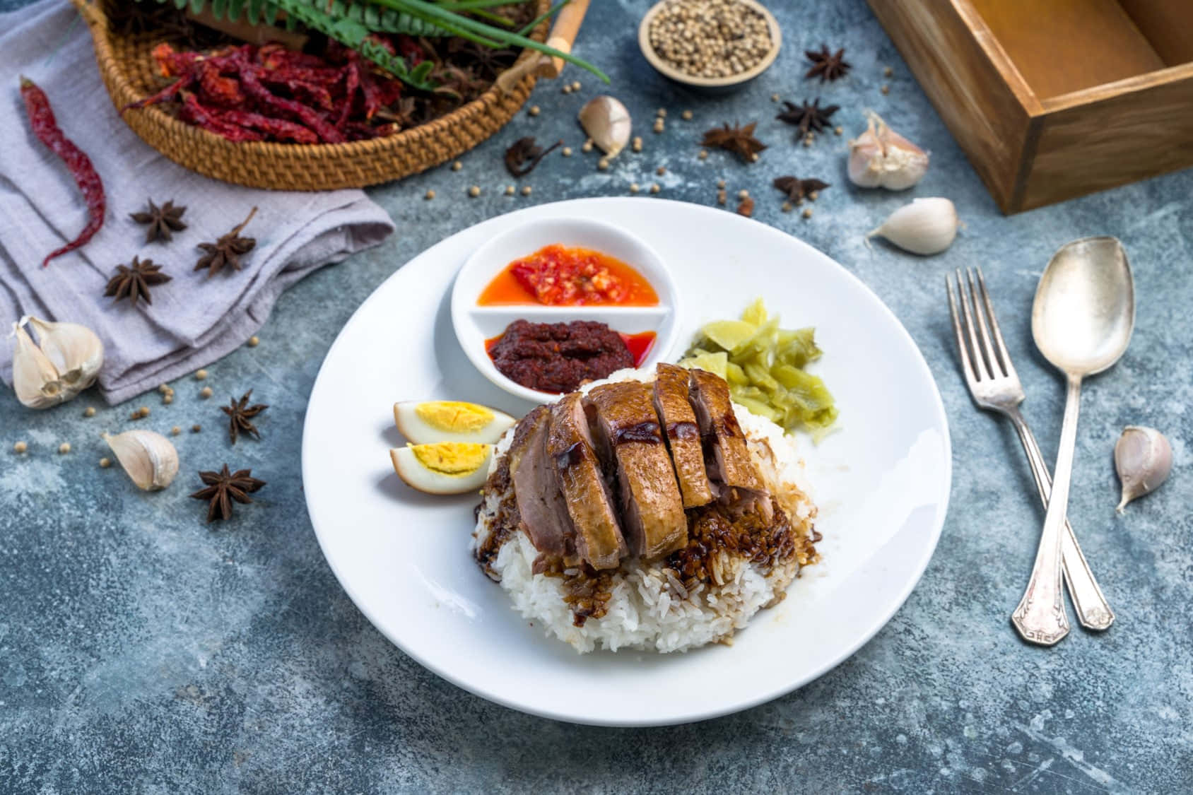 Food Photography 桌布 壁紙 — 一盤切片烤鴨飯，搭配水煮蛋、醃菜和參巴醬，周圍環繞香料，置於有紋理的藍色表面上。