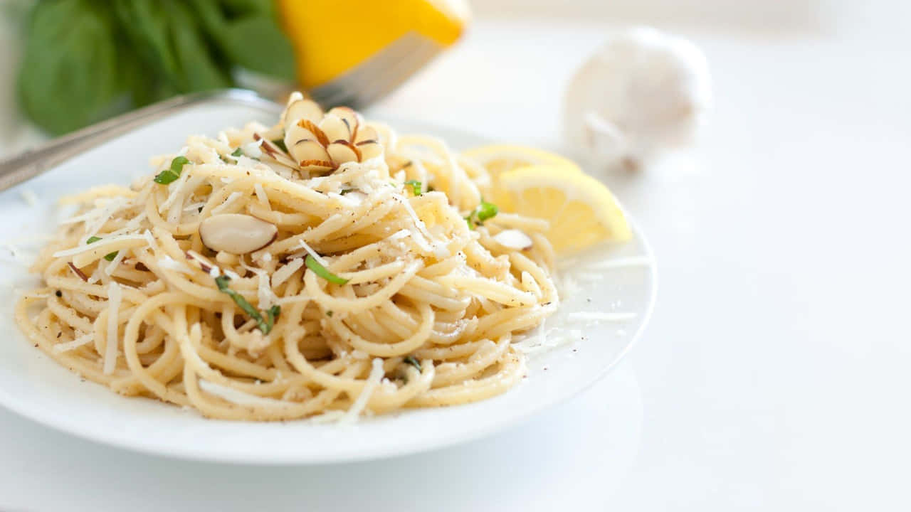 720p Pasta 桌布 壁紙 — 一盤奶油義大利麵，上面放有烤杏仁、新鮮香草和磨碎起司，配以檸檬角、大蒜和菠菜，背景柔和模糊。