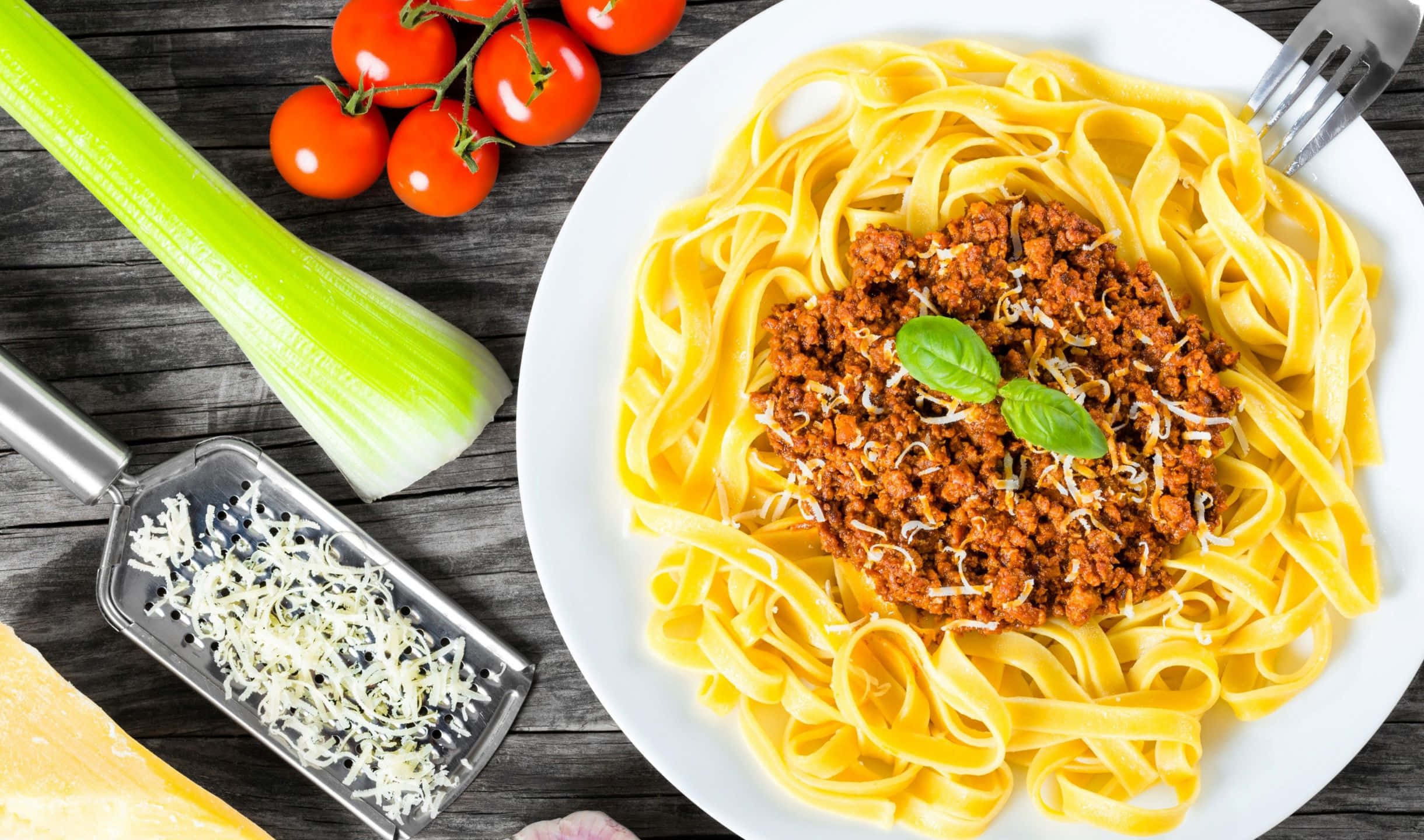 2440x1440 Pasta 桌布 壁紙 — 一盤寬條麵配上濃郁肉醬，以羅勒和碎芝士點綴，旁邊有新鮮番茄和芹菜，放在質樸的木桌上。