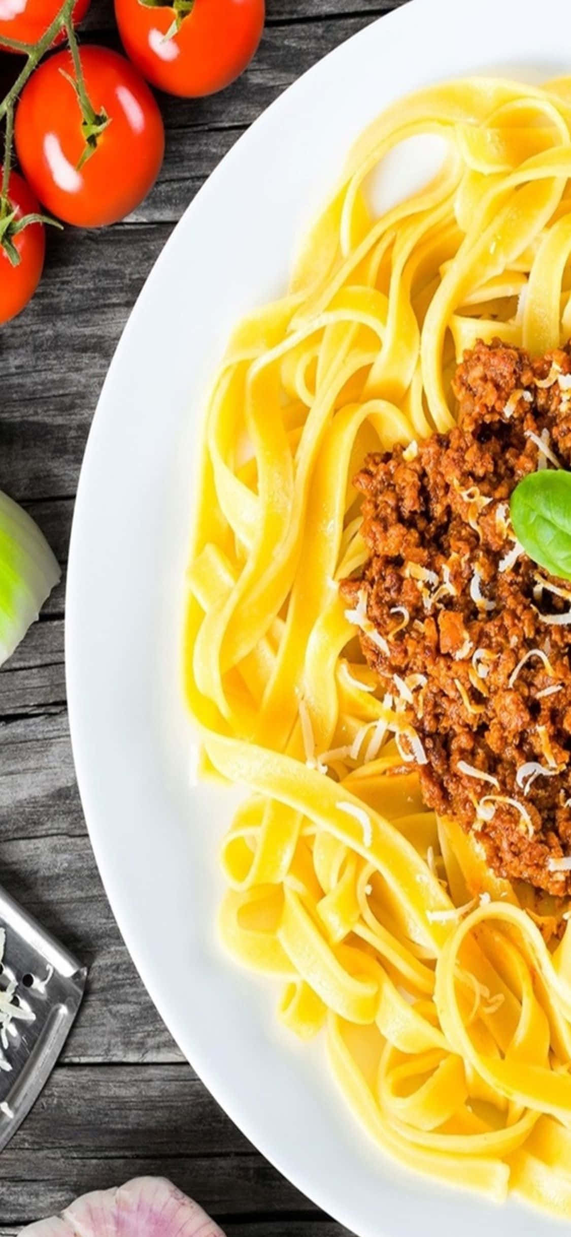iPhone X Pasta 桌布 壁紙 — 一盤寬條麵配上濃郁肉醬和磨碎起司，以羅勒裝飾，放在質樸的木桌上。