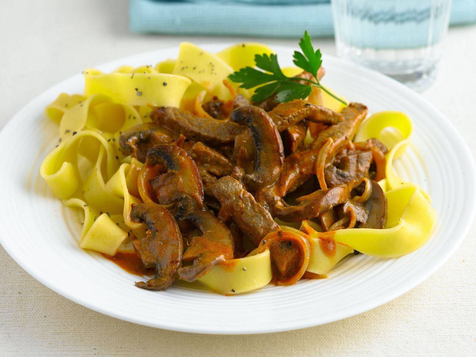 Pasta 桌布 壁紙 — 一盤寬條黃色義大利麵，上面鋪滿炒蘑菇和洋蔥，搭配濃郁的棕色醬汁，並以新鮮歐芹點綴。