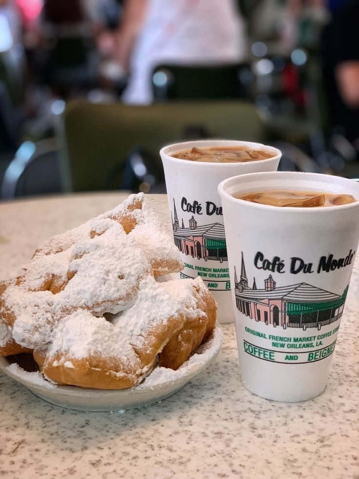 Blue Bayou 桌布 壁紙 — 一盤撒滿糖粉的貝涅餅旁邊有兩杯 Café Du Monde 的冰咖啡，放在有斑點的檯面上。