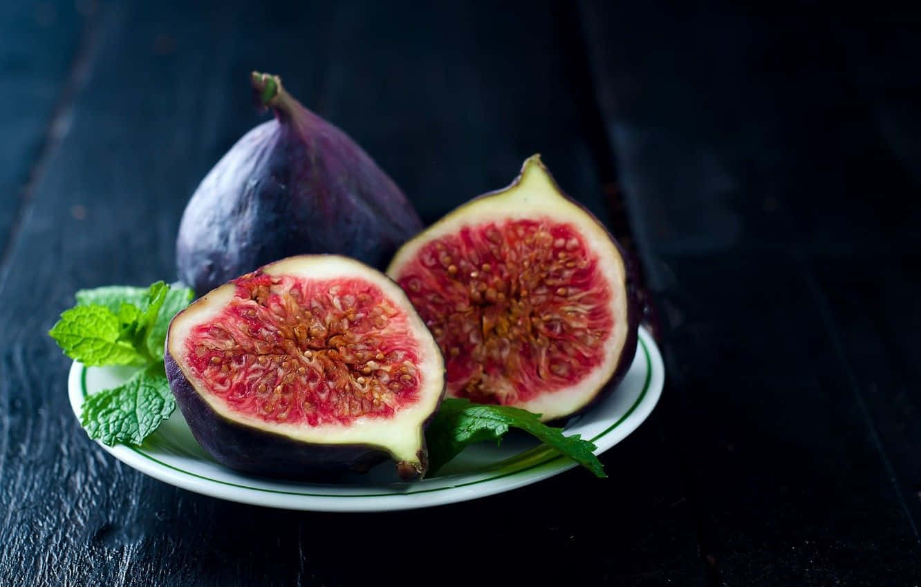 Purple Figs 桌布 壁紙 — 一盤新鮮無花果切開，露出多汁的紅色果肉和綠色薄荷葉，放在深色木桌上。