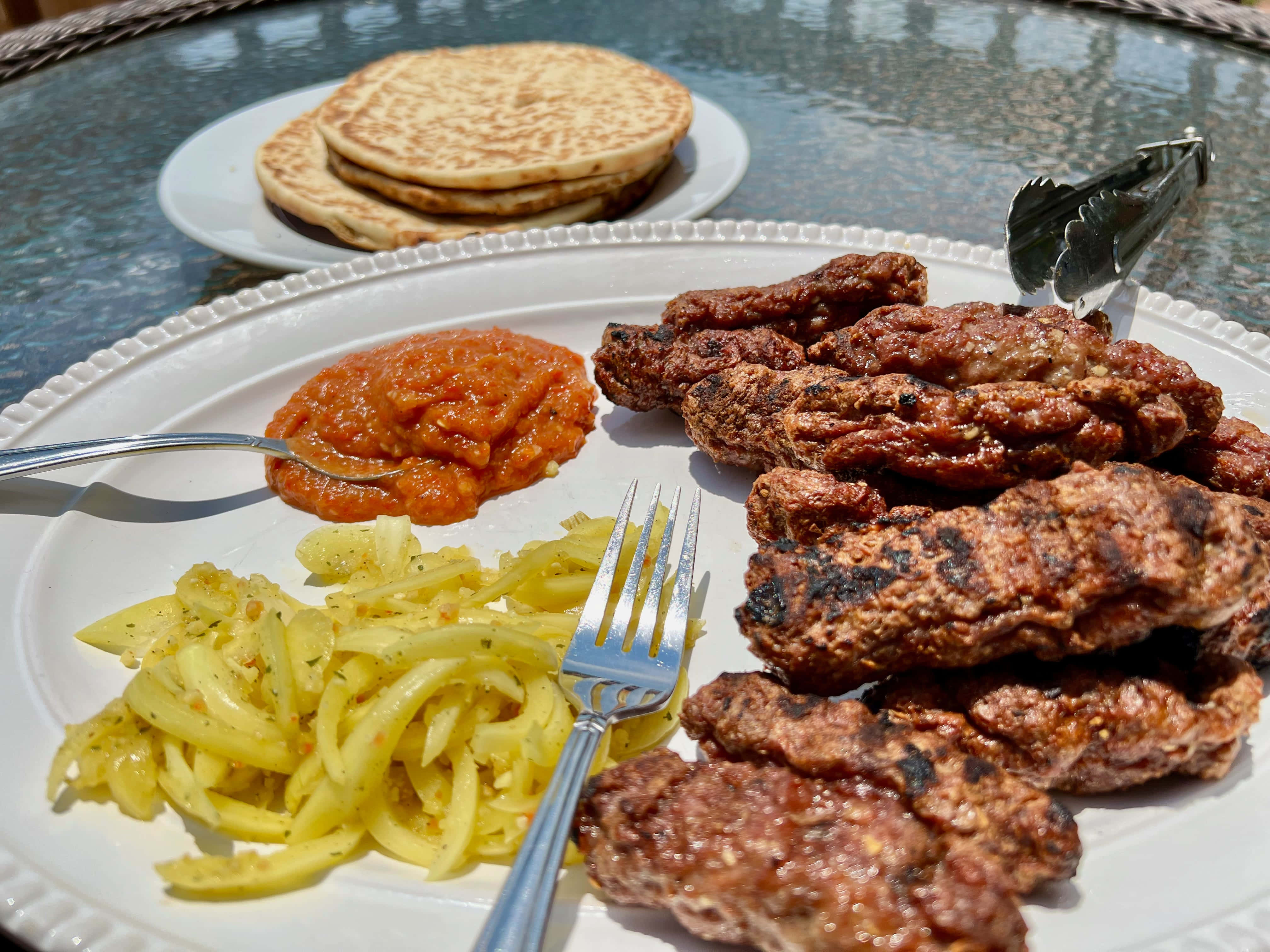meat 桌布 壁紙 — 一盤烤串、番茄醬和螺旋形黃色櫛瓜，旁邊是疊起的煎餅，放在戶外的玻璃桌上。