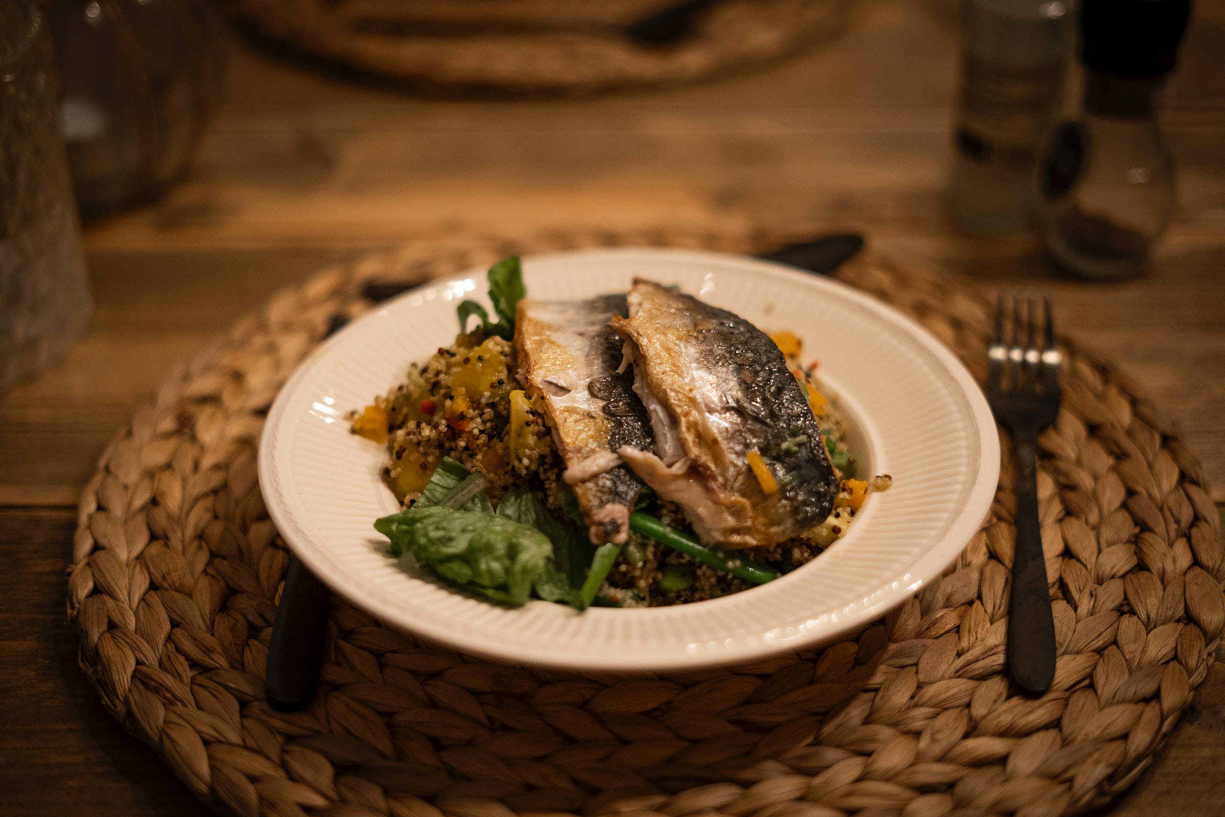 Food Photography 桌布 壁紙 — 一盤烤魚搭配藜麥和青菜，溫暖地照亮在木桌上編織的餐墊上。