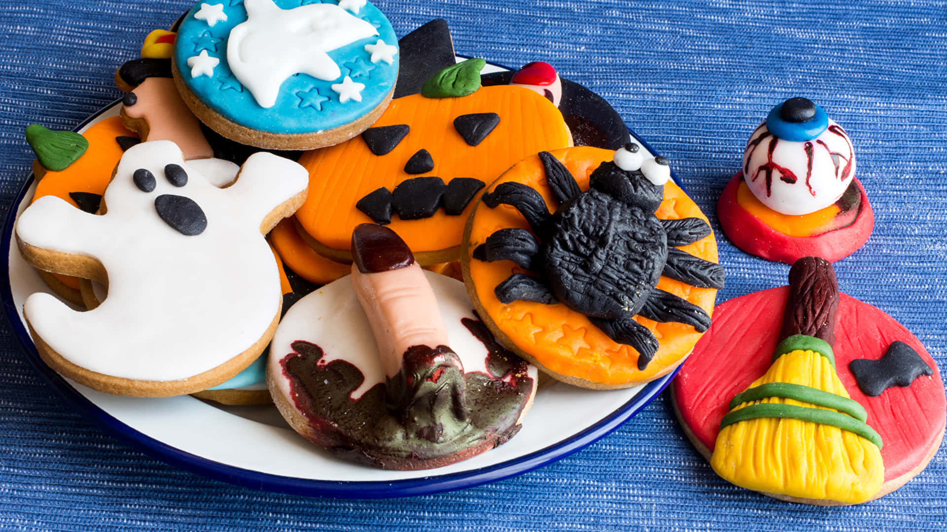Halloween Treats 桌布 壁紙 — 一盤精心裝飾的萬聖節餅乾，形狀有幽靈、南瓜、蜘蛛和女巫掃帚，放在藍色布上。