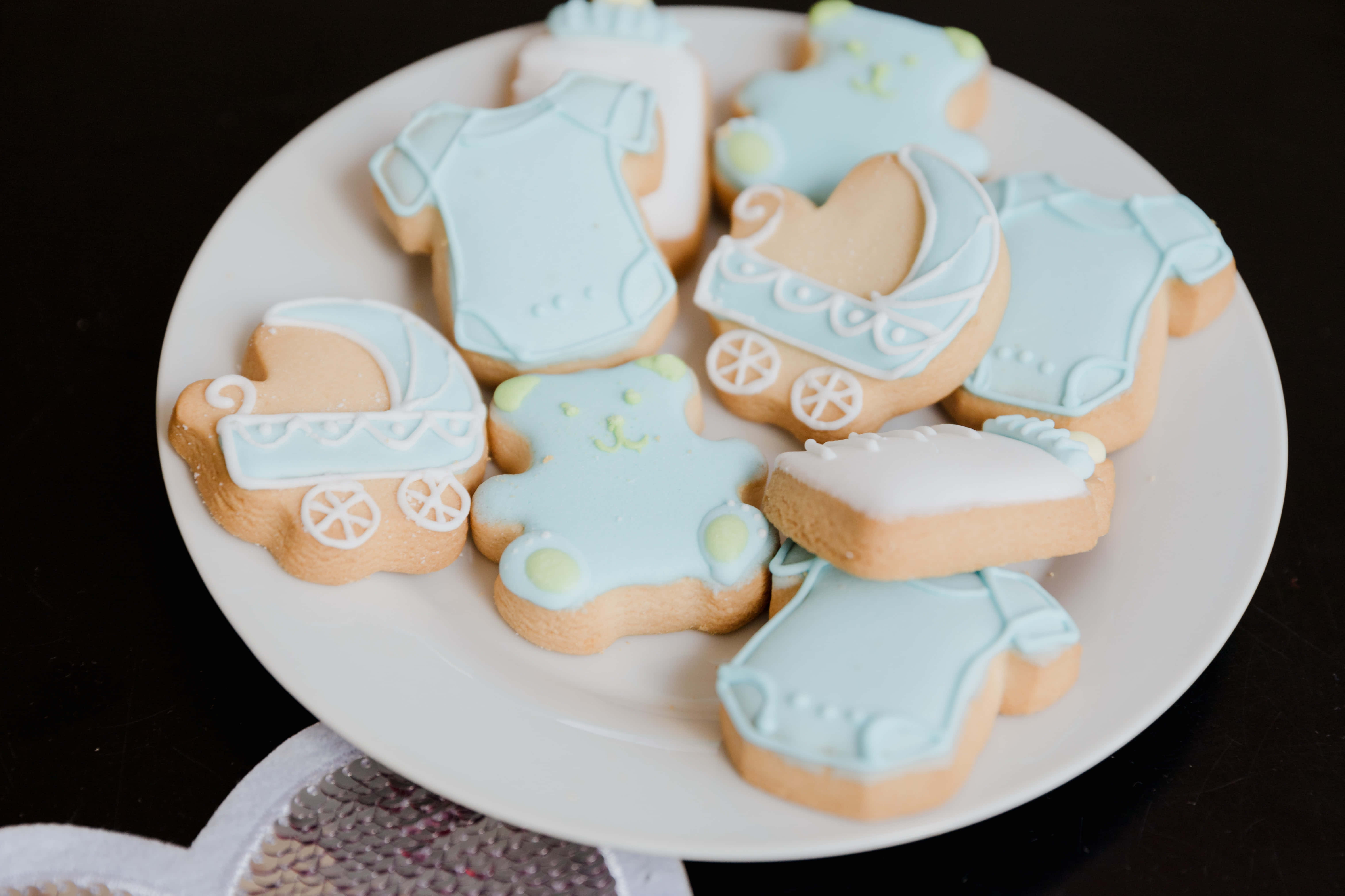 Baby Shower 桌布 壁紙 — 一盤精緻的嬰兒主題糖霜餅乾，造型為連體衣和嬰兒車，以柔和的藍色和白色糖霜裝飾，排列在深色表面上。