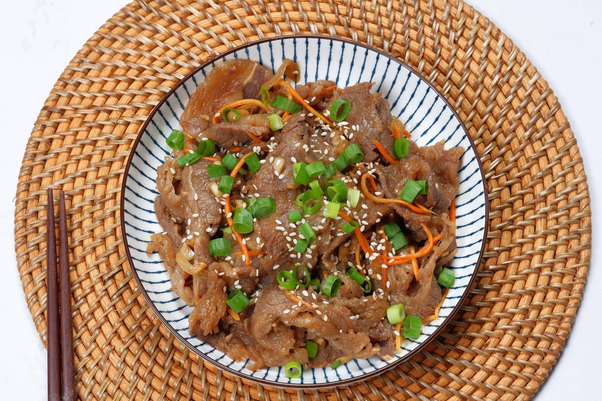Carrot 桌布 壁紙 — 一盤美味牛肉，上面撒有芝麻、青蔥和蘿蔔絲，放在編織墊上，旁邊有筷子。