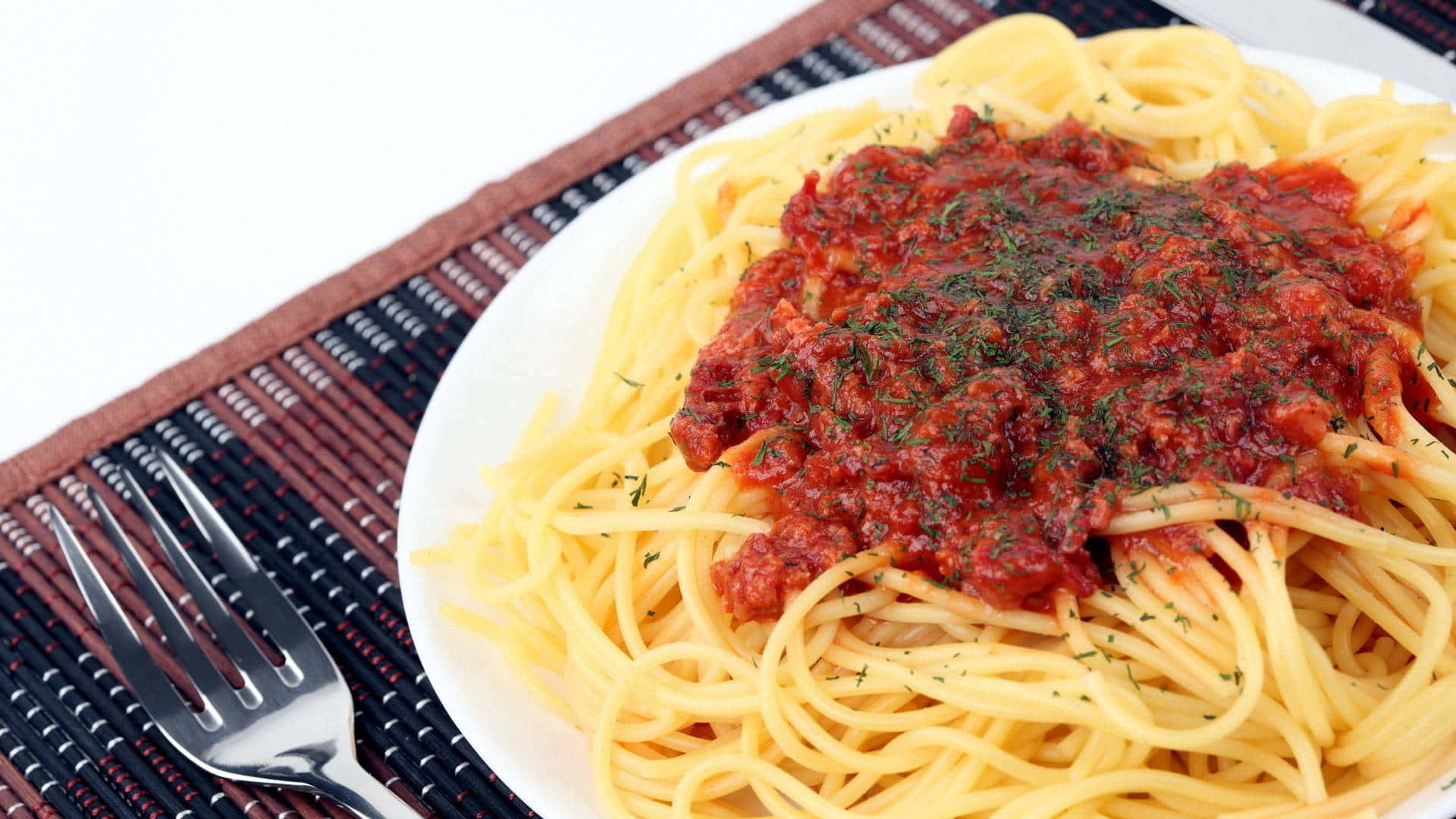 Spaghetti 桌布 壁紙 — 一盤義大利麵配上濃郁番茄醬和新鮮香草，放在編織餐墊上，旁邊有一把叉子。