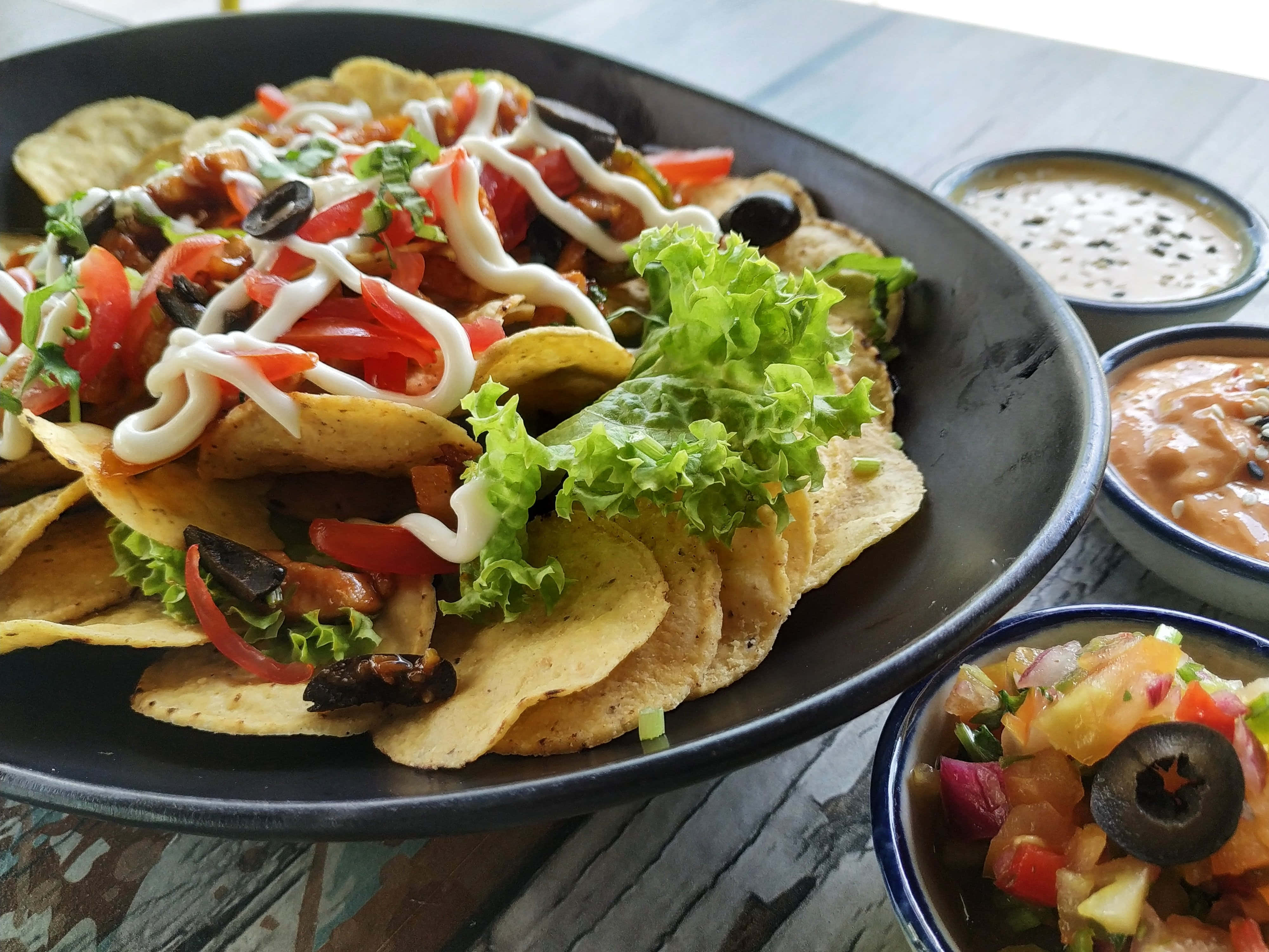 Mexican 桌布 壁紙 — 一盤色彩繽紛的滿載玉米片，上面鋪滿新鮮蔬菜、酸奶油和生菜，旁邊有三款蘸醬，放在質樸的木桌上。