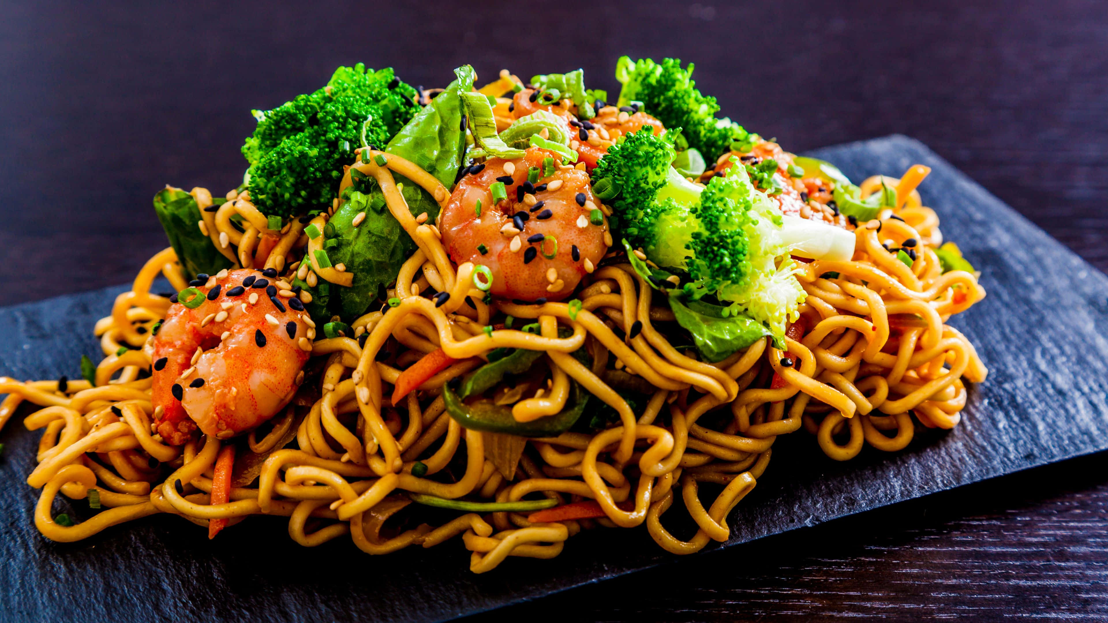 Noodles 桌布 壁紙 — 一盤色彩繽紛的炒金黃麵條，配上蝦仁、花椰菜和芝麻，高高堆在深色石板盤上。