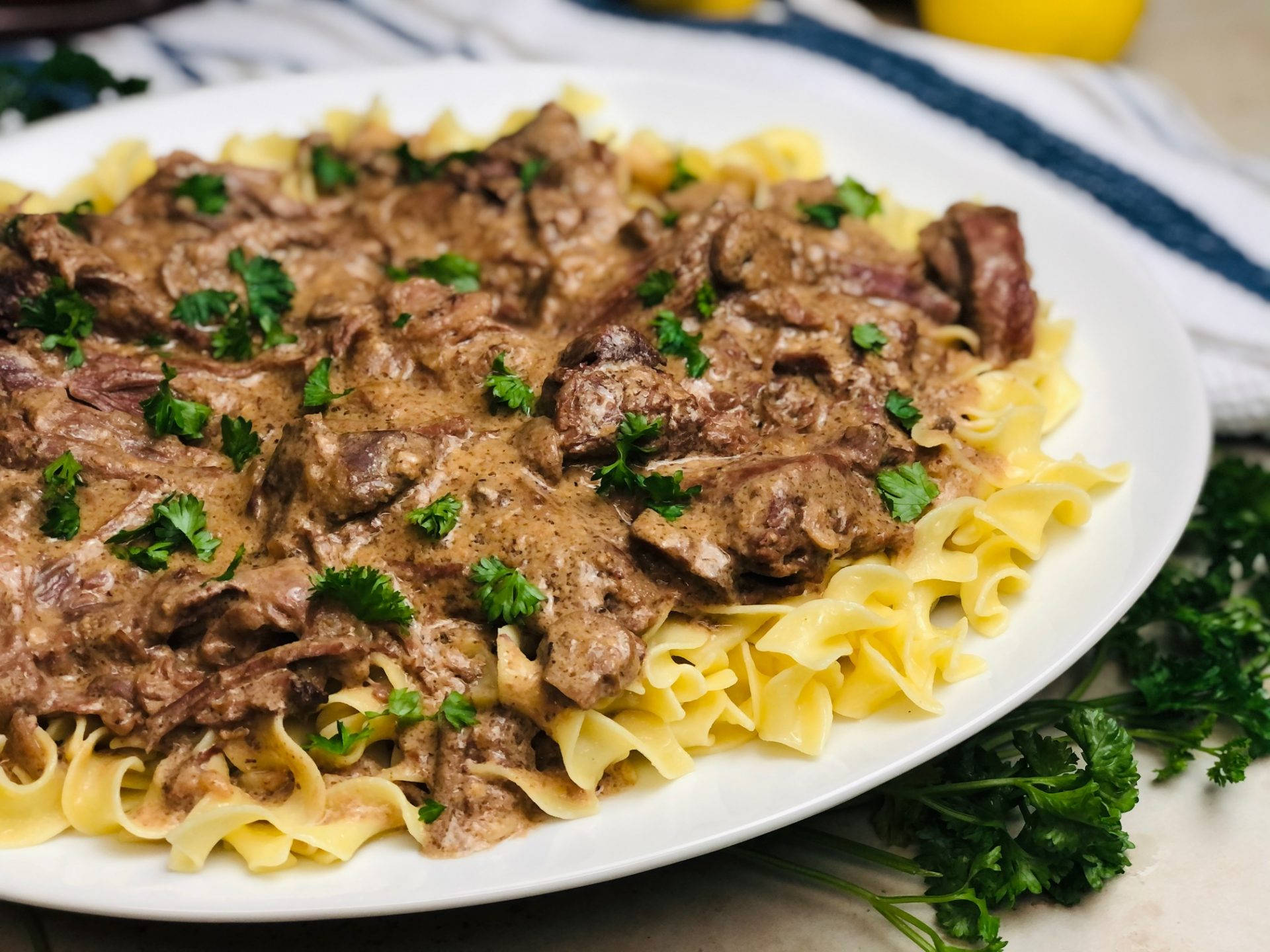 Food Photography 桌布 壁紙 — 一盤豐富的牛肉斯特羅加諾夫配雞蛋麵，上面撒上新鮮歐芹，放在條紋布上。