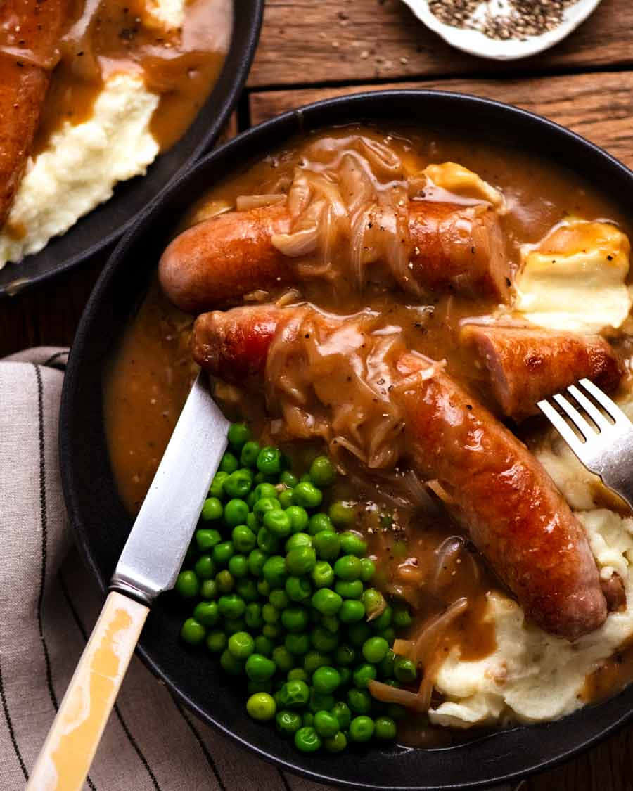 Sausage 桌布 壁紙 — 一盤豐盛的香腸，浸在濃郁肉汁和焦糖洋蔥中，配上馬鈴薯泥和鮮綠豌豆，放在質樸的木桌上。