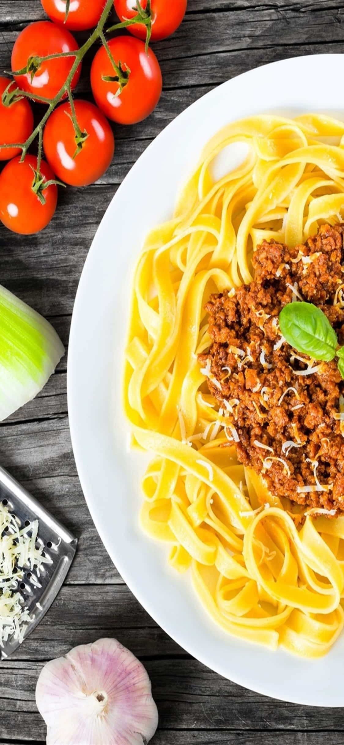 iPhone X Pasta 桌布 壁紙 — 一盤配有濃郁肉醬和新鮮羅勒的義大利寬麵條，旁邊擺放著鮮豔的櫻桃番茄、大蒜和磨碎的起司，置於鄉村風木桌上。