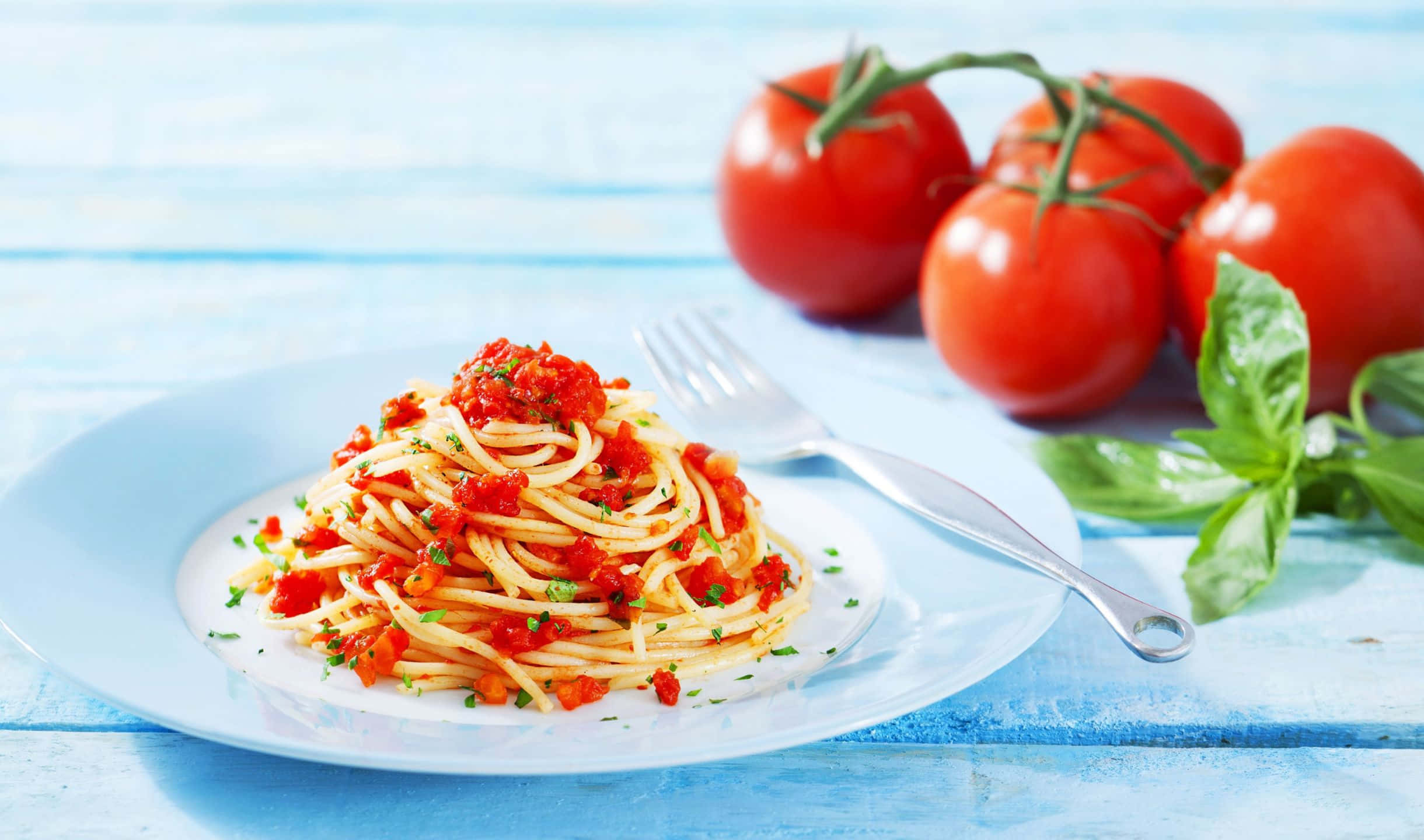 2440x1440 Pasta 桌布 壁紙 — 一盤配有番茄醬和羅勒的義大利麵放在藍色木桌上，旁邊是新鮮番茄和羅勒。