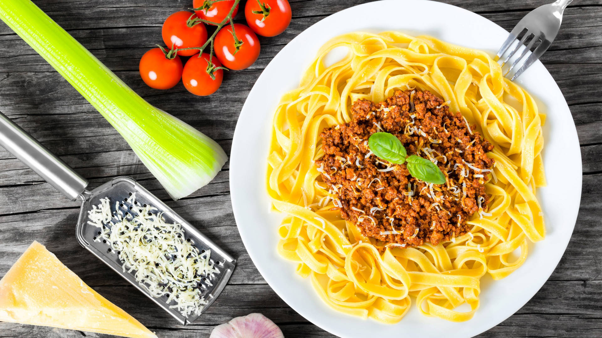 Pasta 桌布 壁紙 — 一盤配有肉醬、羅勒和碎起司的寬條麵，旁邊放著新鮮番茄、芹菜和大蒜，放在質樸的木桌上。