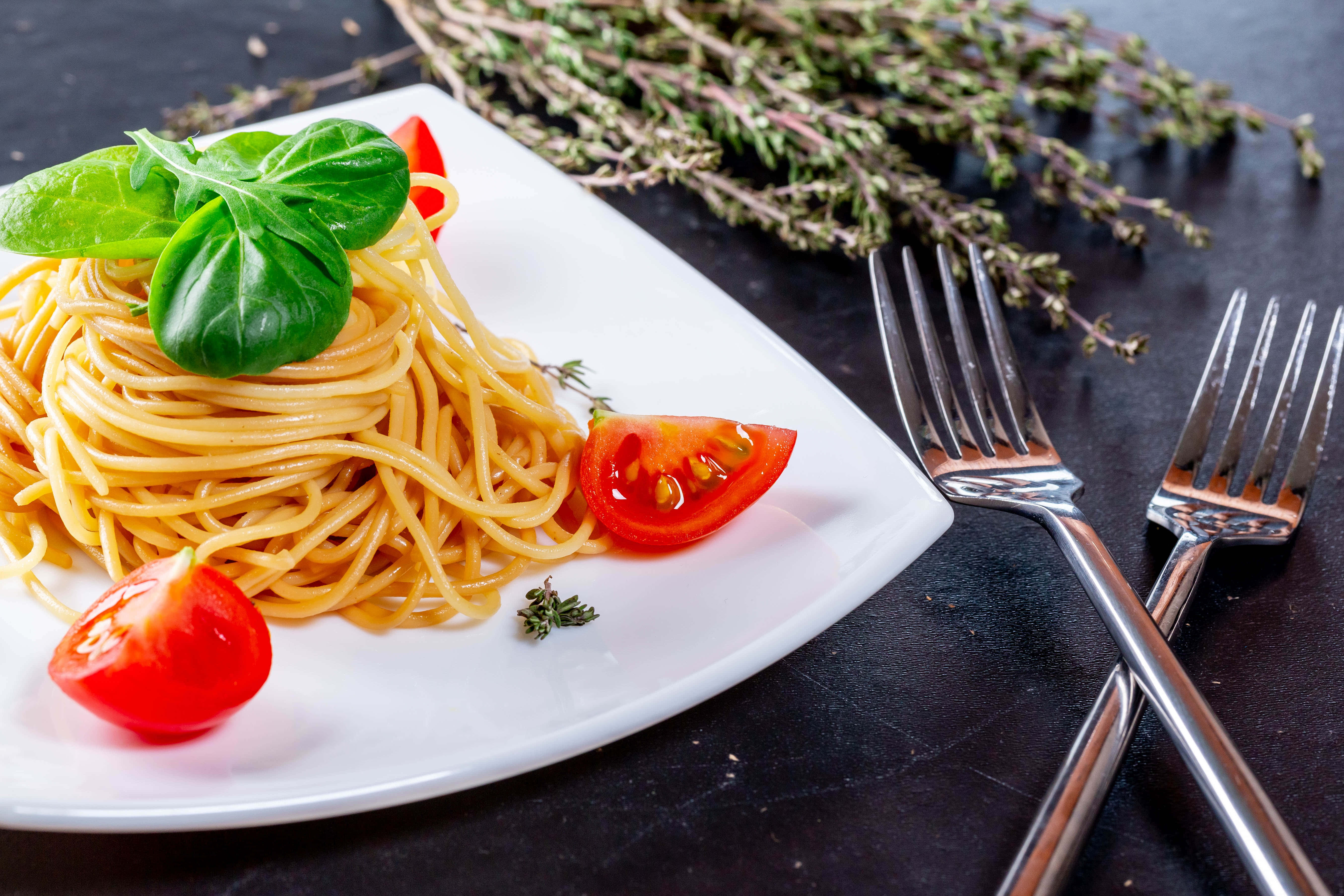 4K Pasta 桌布 壁紙 — 一盤金黃色義大利麵，上面放上新鮮羅勒和櫻桃番茄，放在深色鄉村風格的桌子上，配有叉子。