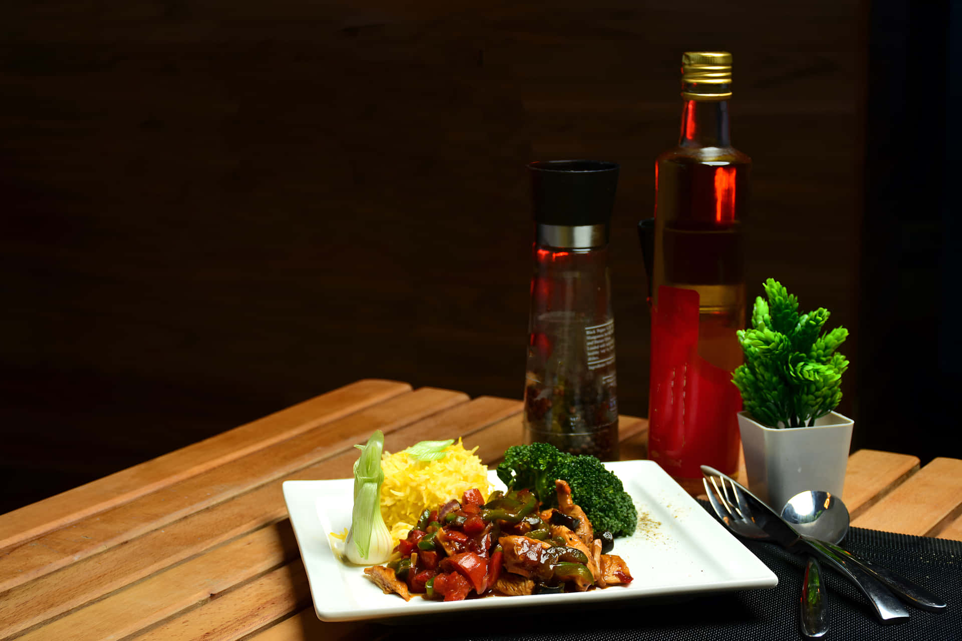 Chicken Chilli Dry with rice on a plate 壁紙 — 一盤雞肉炒西蘭花和黃色米飯的餐點放在木桌上，旁邊有一瓶紅色液體和一株小綠色植物。