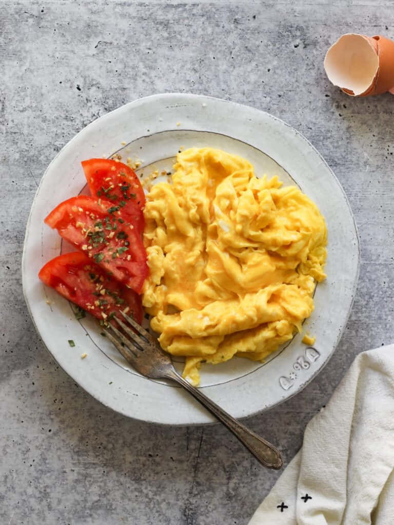 Scrambled Egg 桌布 壁紙 — 一盤鬆軟炒蛋和新鮮番茄片，用香草調味，旁邊放著蛋殼，置於紋理灰色表面上。