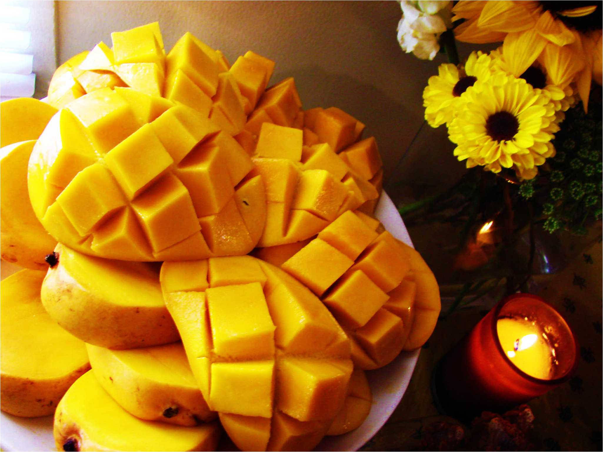 Mango 桌布 壁紙 — 一盤鮮豔的芒果丁放在向日葵花瓶和發光蠟燭旁邊，在溫暖的環境光中。