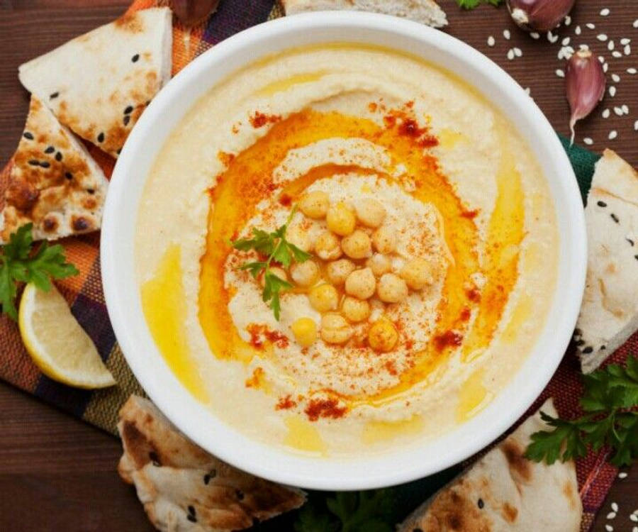 Hummus 桌布 壁紙 — 一碗 creamy 的鷹嘴豆泥，飾以橄欖油、紅椒粉、鷹嘴豆和歐芹，周圍有 pita 麵包、檸檬和蒜瓣。