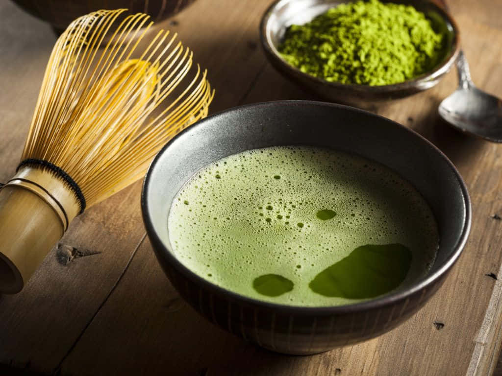 Matcha 桌布 壁紙 — 一碗充滿泡沫的鮮綠色抹茶旁邊放著竹筅和粉末，置於質樸的木桌上。