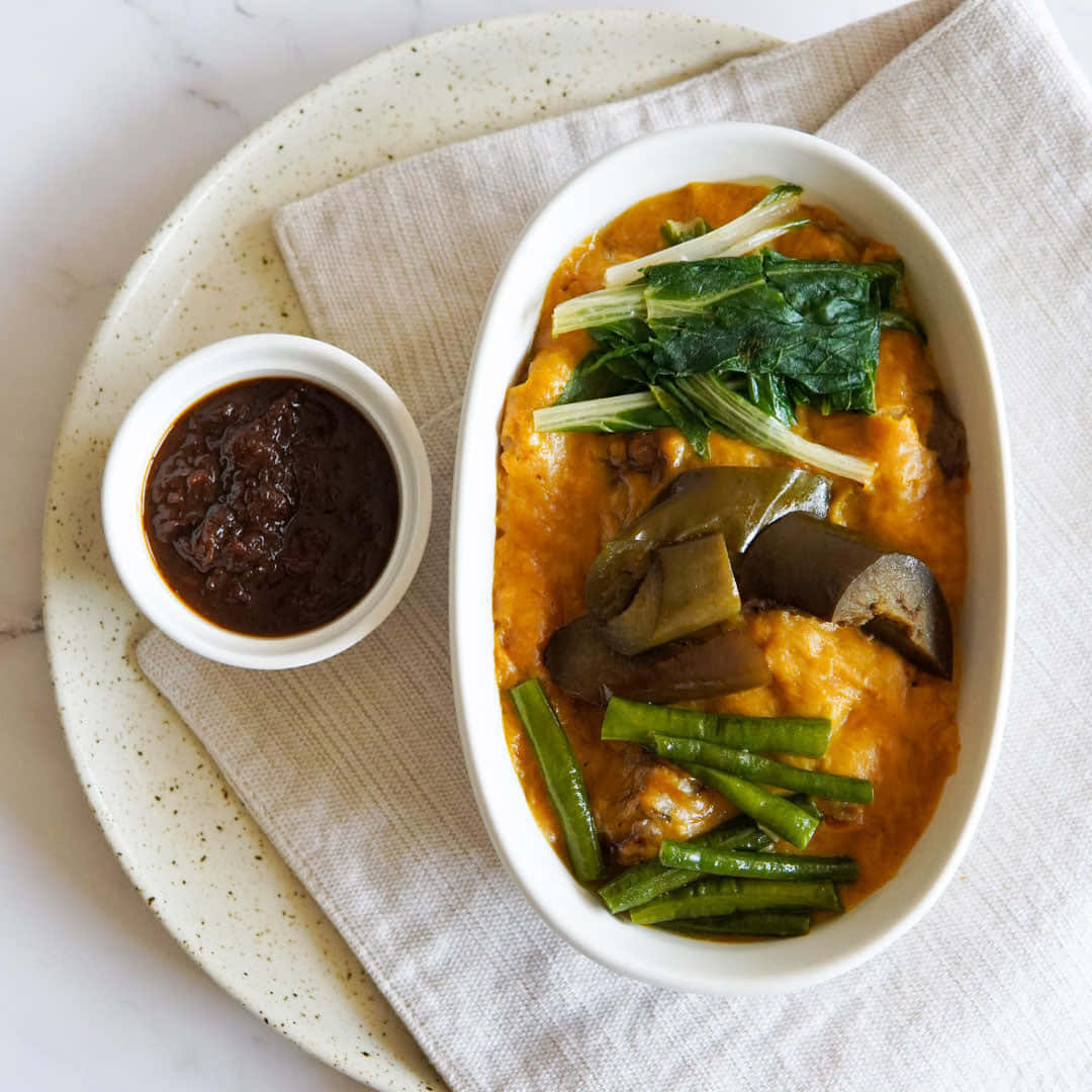 meat 桌布 壁紙 — 一碗充滿活力的橙色咖哩，搭配茄子、青豆和菠菜，盛在白色碗中，旁邊有深色醬汁，放在斑點盤上。