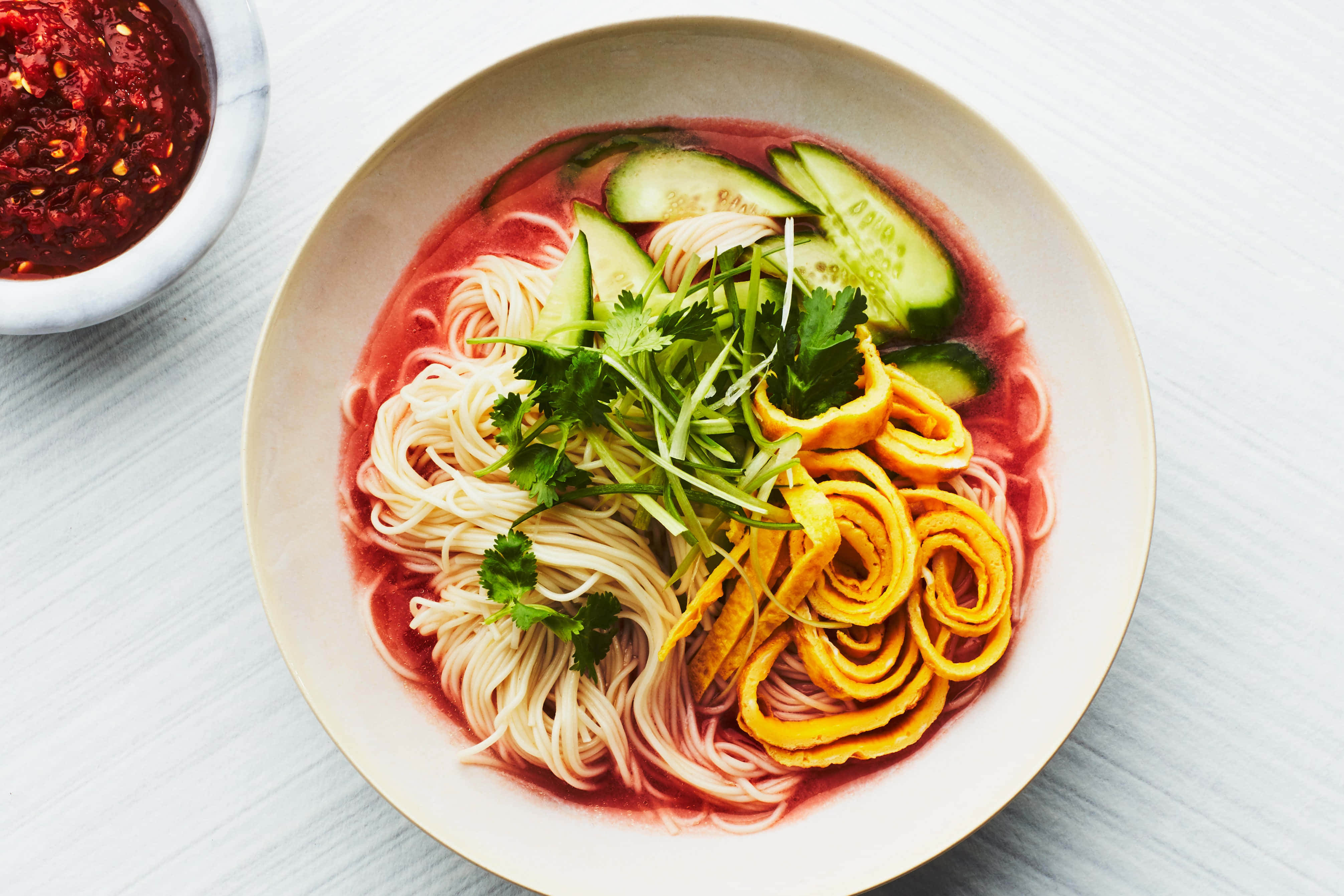 Food Photography 桌布 壁紙 — 一碗充滿活力的粉紅色湯麵，上面放有黃瓜、香草和黃色春捲，旁邊是辣味蘸醬。