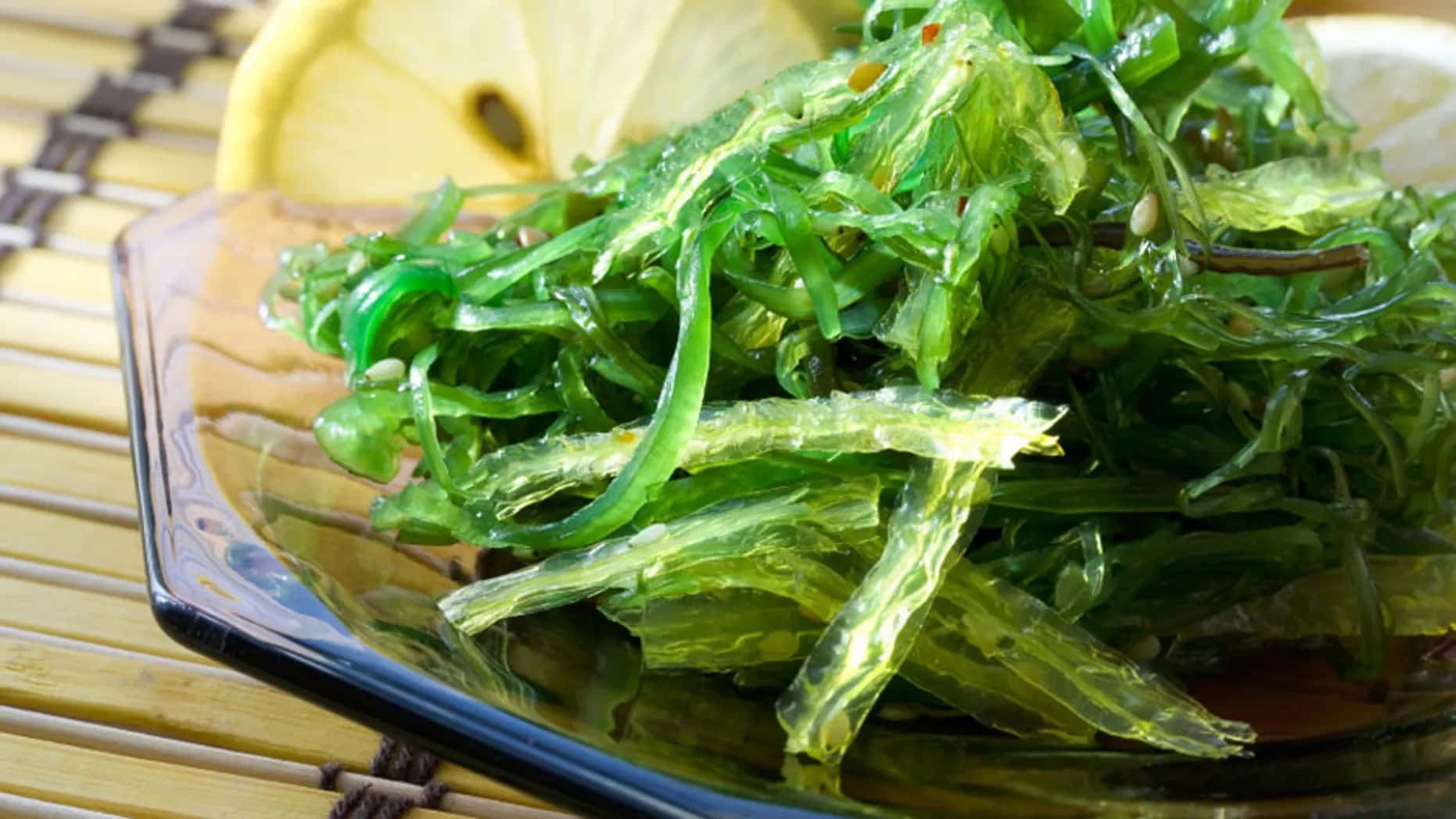 Sea Weed 桌布 壁紙 — 一碗充滿活力的綠色海藻沙拉在深色玻璃碗中閃閃發光，旁邊是竹蓆上的檸檬片。