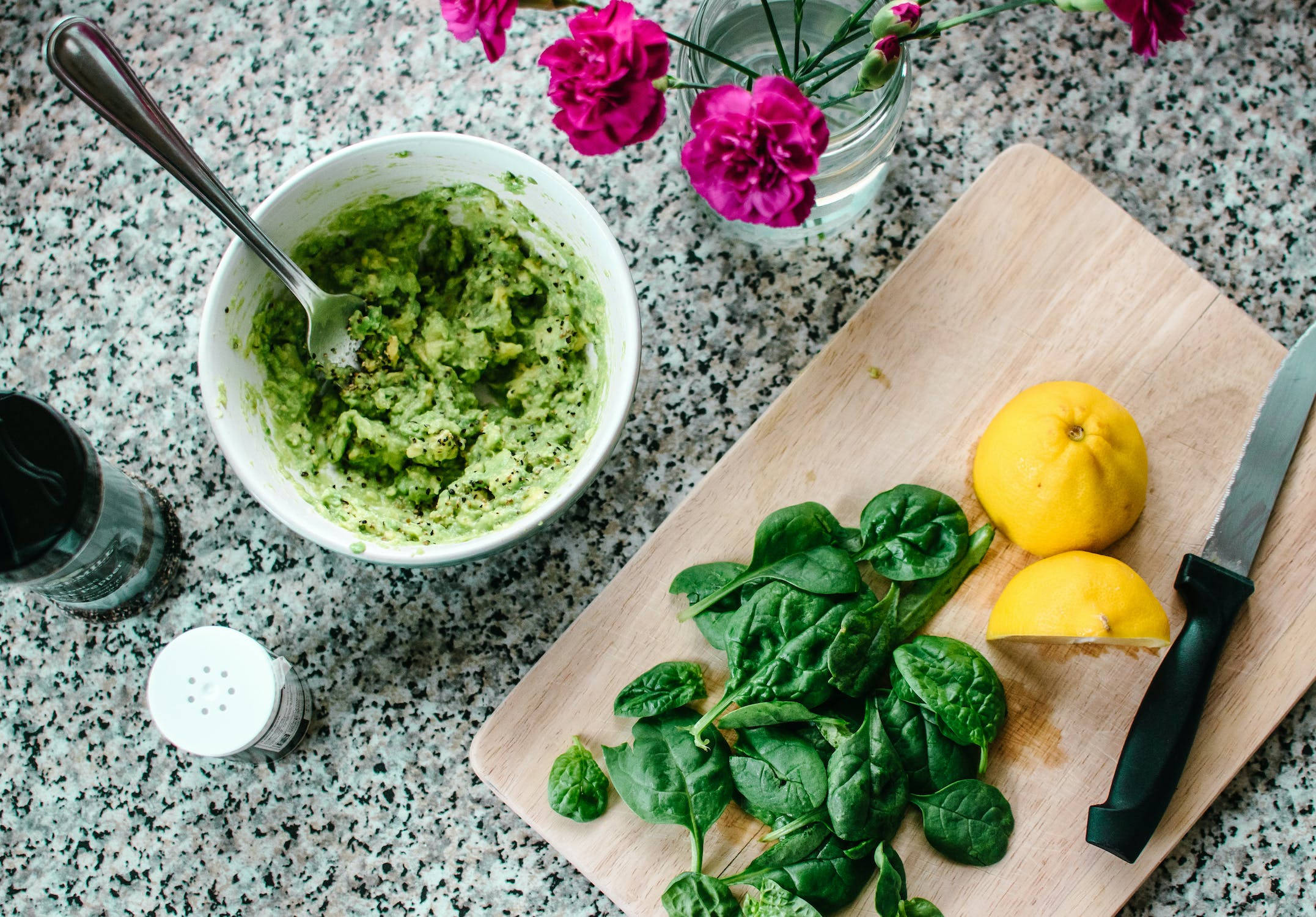 Healthy Food 桌布 壁紙 — 一碗充滿活力的綠色酪梨醬放在有斑點的檯面上，旁邊是新鮮菠菜、檸檬和一瓶紫紅色康乃馨。